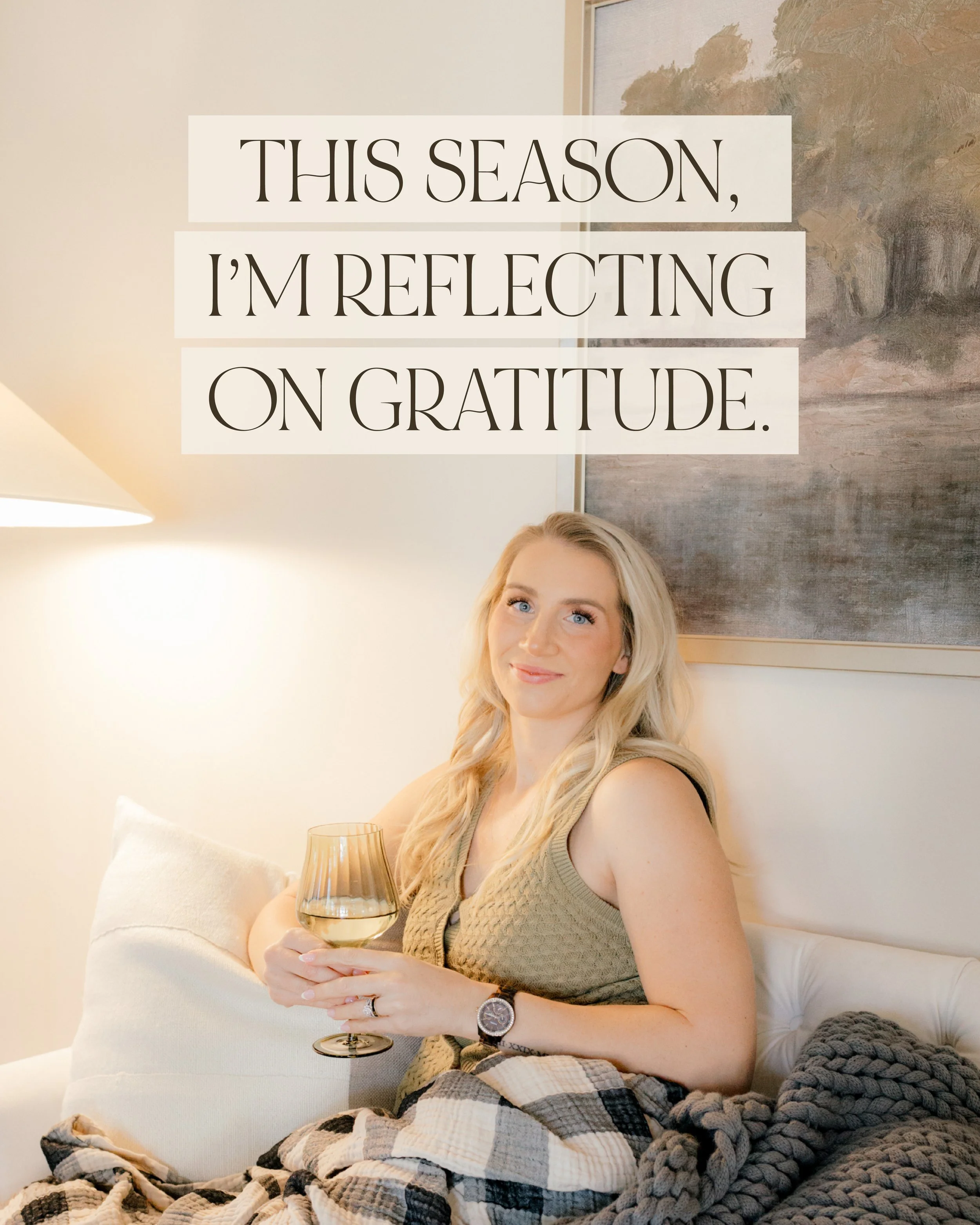 A woman with blonde hair sitting on a white sofa. She is holding a glass of white wine, wearing a beige knitted sleeveless top and a watch. Behind her, there is a painting and a lamp. Some blankets are draped on the sofa.
