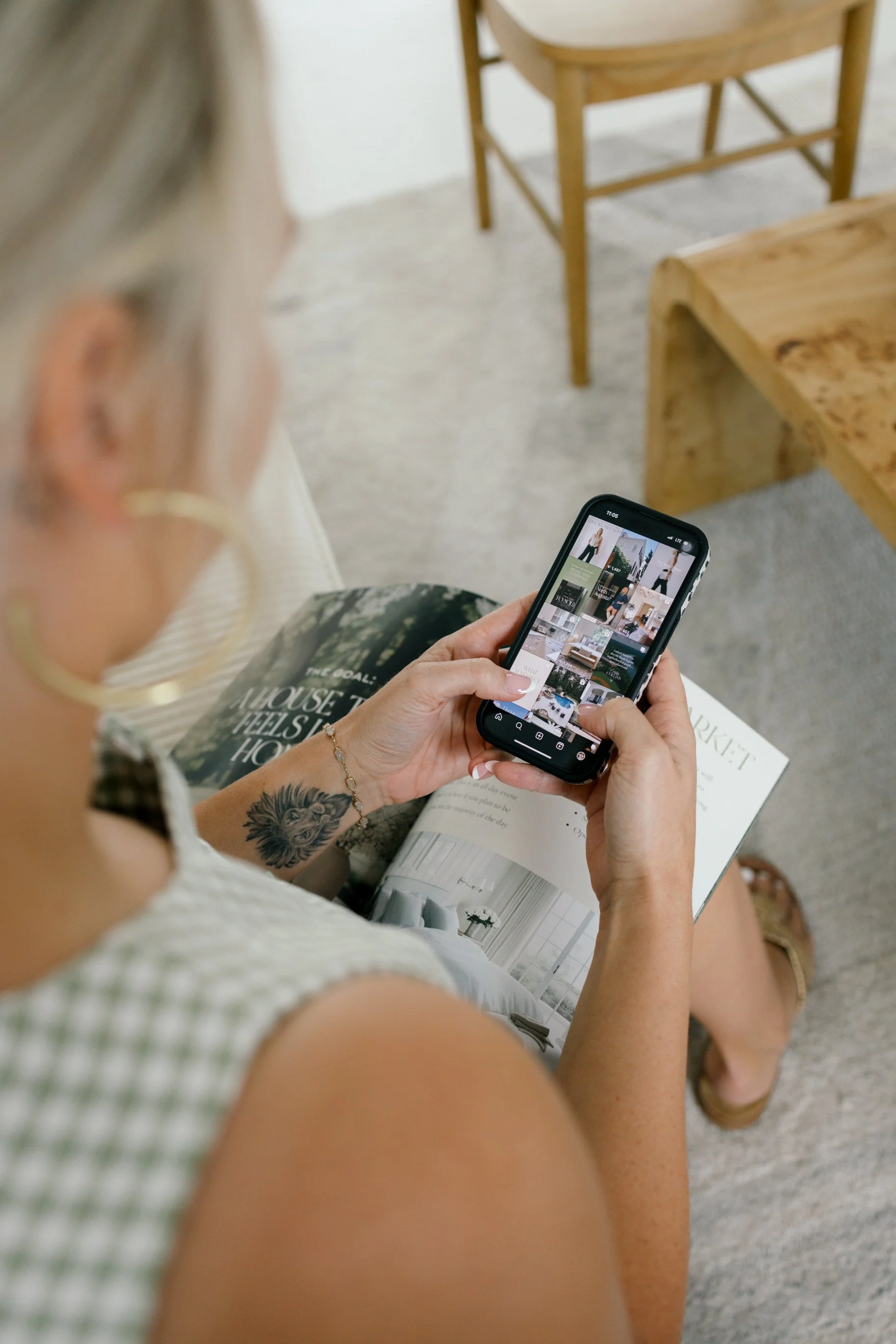 A person with a tattooed arm sitting on a chair, holding a smartphone and browsing a website or app, with an open magazine on their lap.