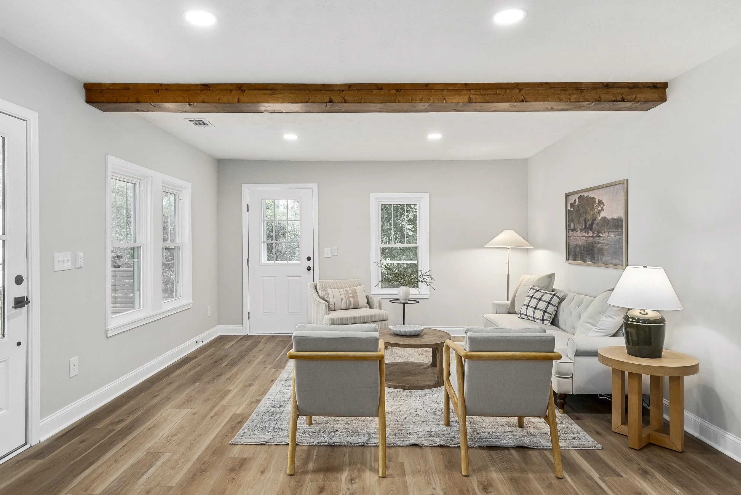 Living room with large windows, white walls, hardwood flooring, and modern furniture, including a sofa, armchairs, a wooden coffee table, and a floor lamp, with a wooden beam on the ceiling.