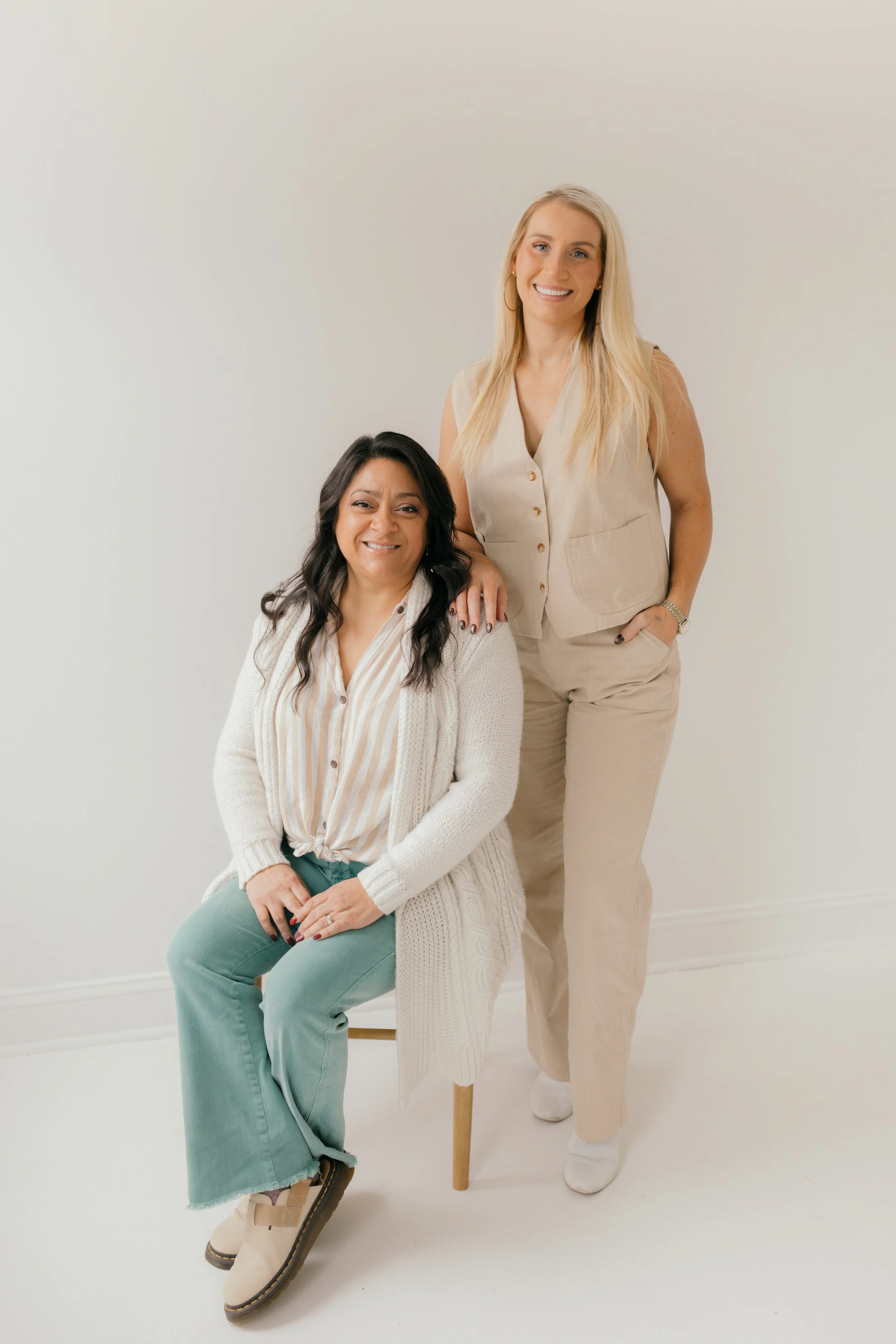 Two women, one sitting on a chair and the other standing behind her on the right, in a bright room with a plain white wall, smiling at the camera. The seated woman has long dark wavy hair, wearing a white cardigan, striped shirt, and teal pants. The standing woman has long blonde hair, wearing a beige sleeveless vest over a light shirt and matching beige pants.