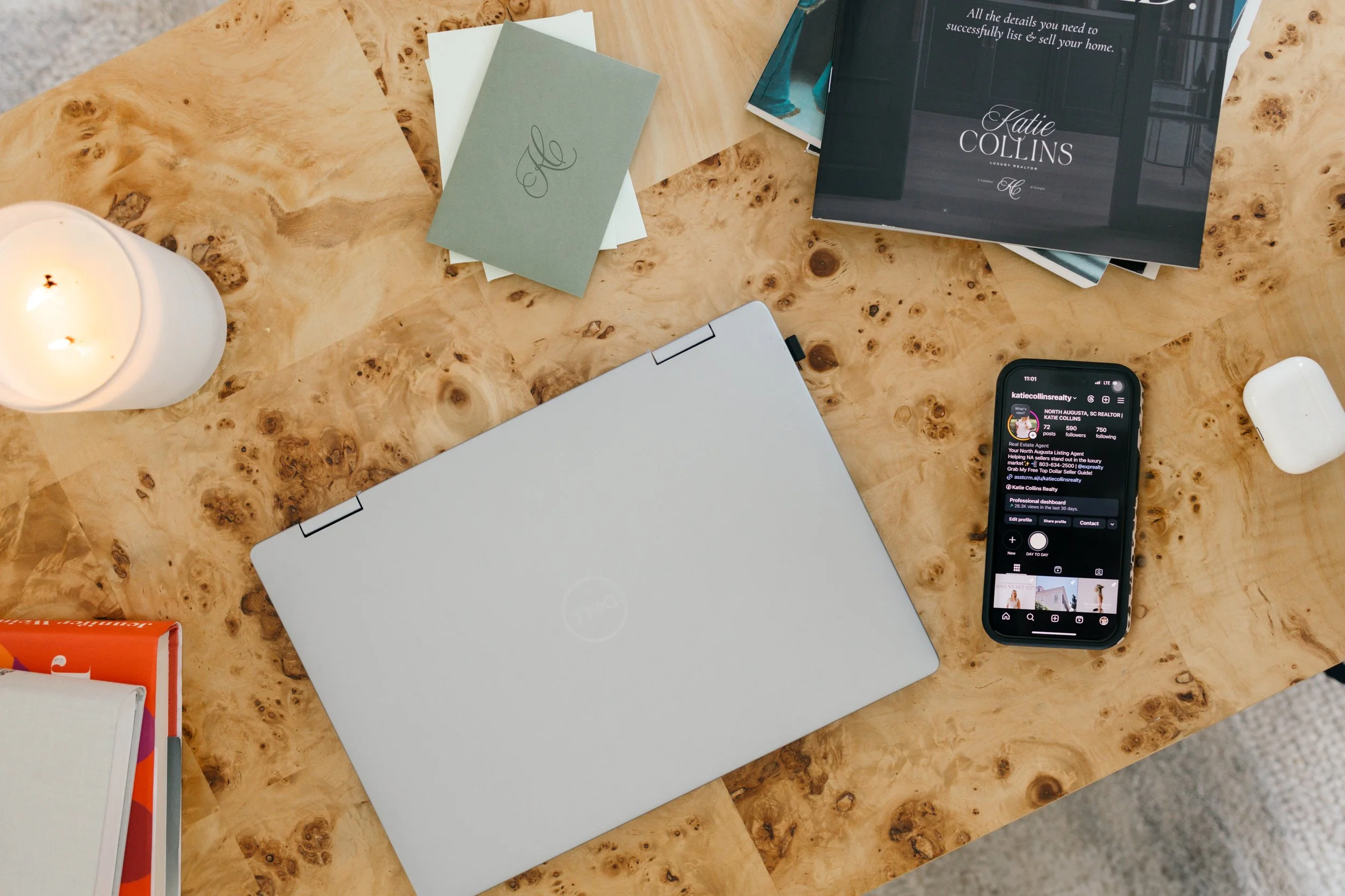 A wooden table with a lit white candle, several papers, a closed laptop, smartphone showing a social media profile, and magazines or pamphlets.