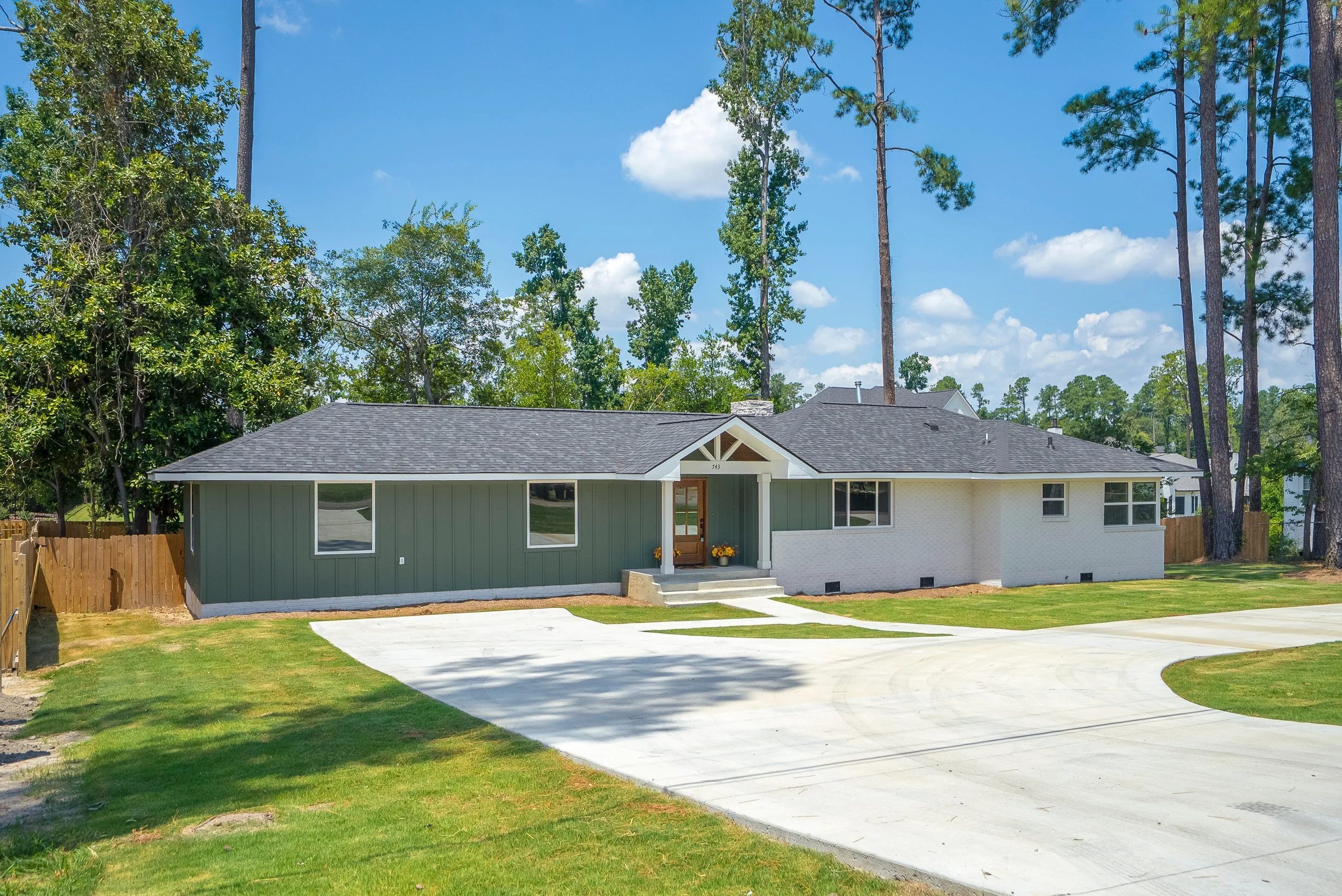 A single-story house with a light green and white exterior, a gable roof, a front porch with steps, a concrete driveway, surrounded by a lawn, and large trees in the background under a blue sky with clouds.