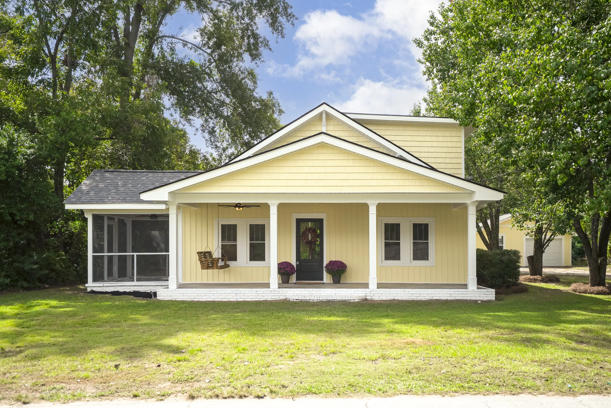 Yellow house with a porch, black front door adorned with a wreath, and two potted pink flowers in black pots. There are two windows with curtains on either side of the door. A wooden porch swing hangs on the left side, and the house is surrounded by green trees and grass under a partly cloudy sky.