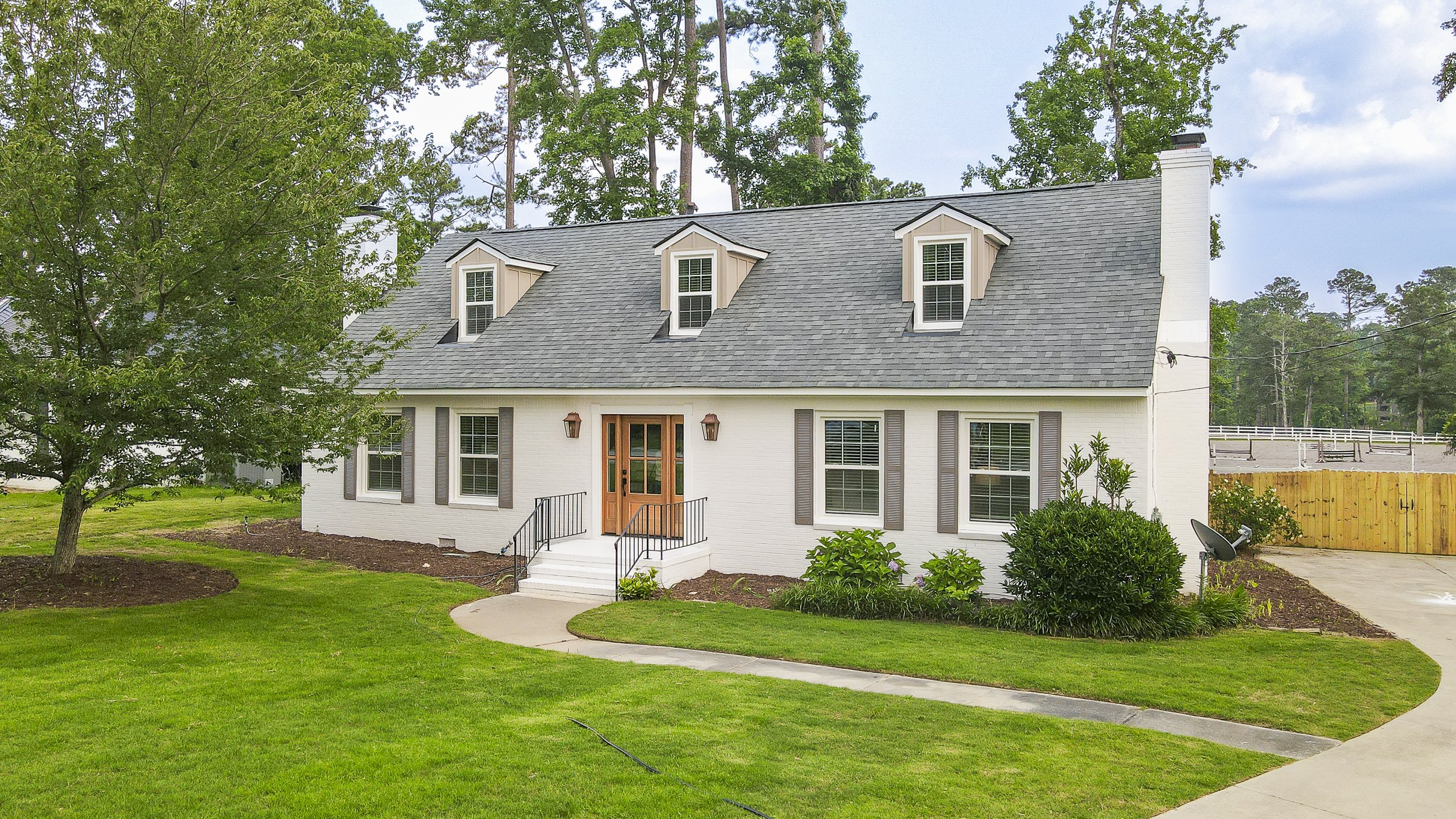 White house with gray roof and multiple windows, surrounded by green lawn and trees, with a concrete walkway leading to the front door.