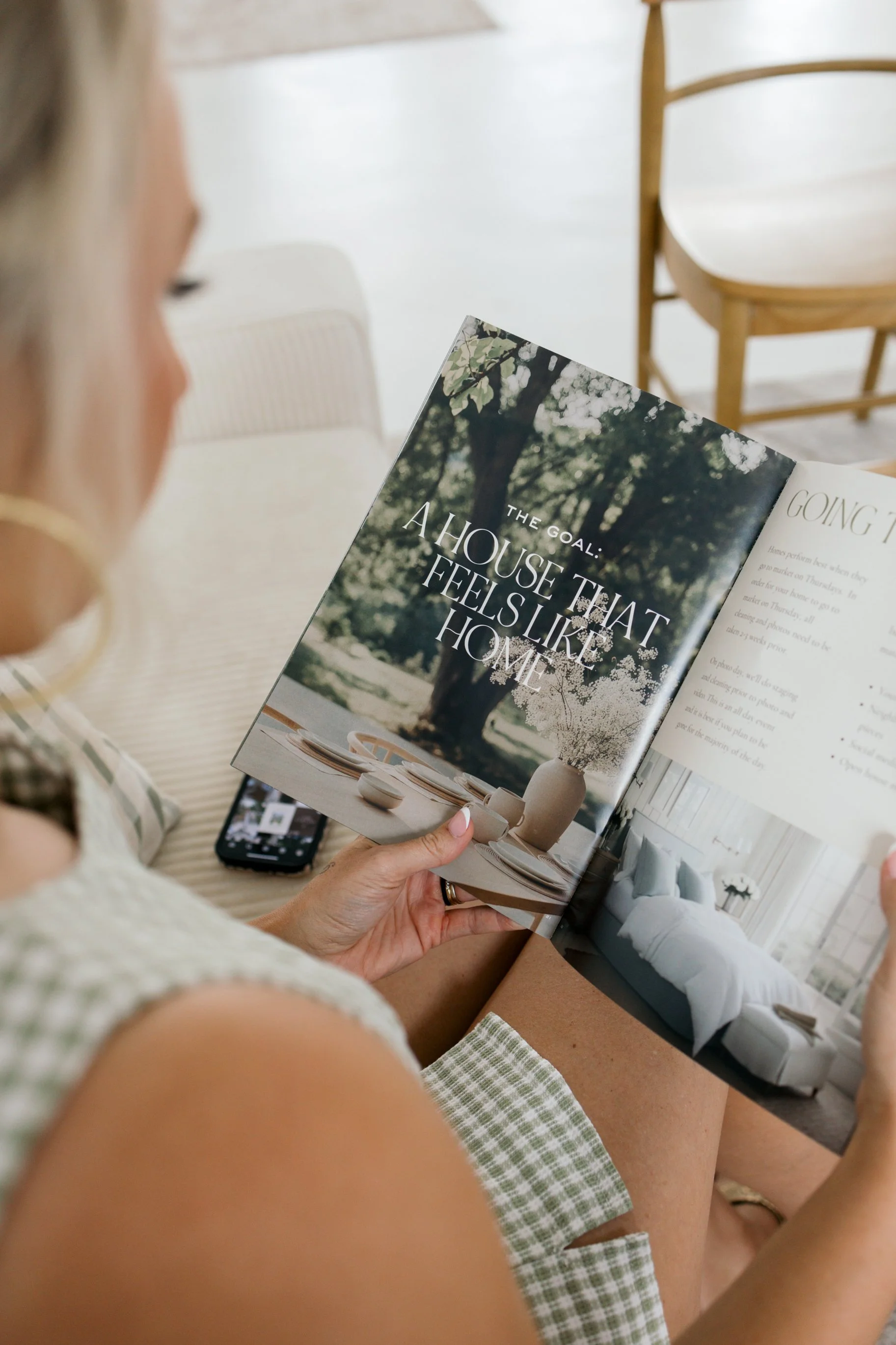 Woman sitting on a beige couch reading a magazine titled "The Goal: The house that feels like home" in a bright, modern living room with wooden chairs and decorative pillows.
