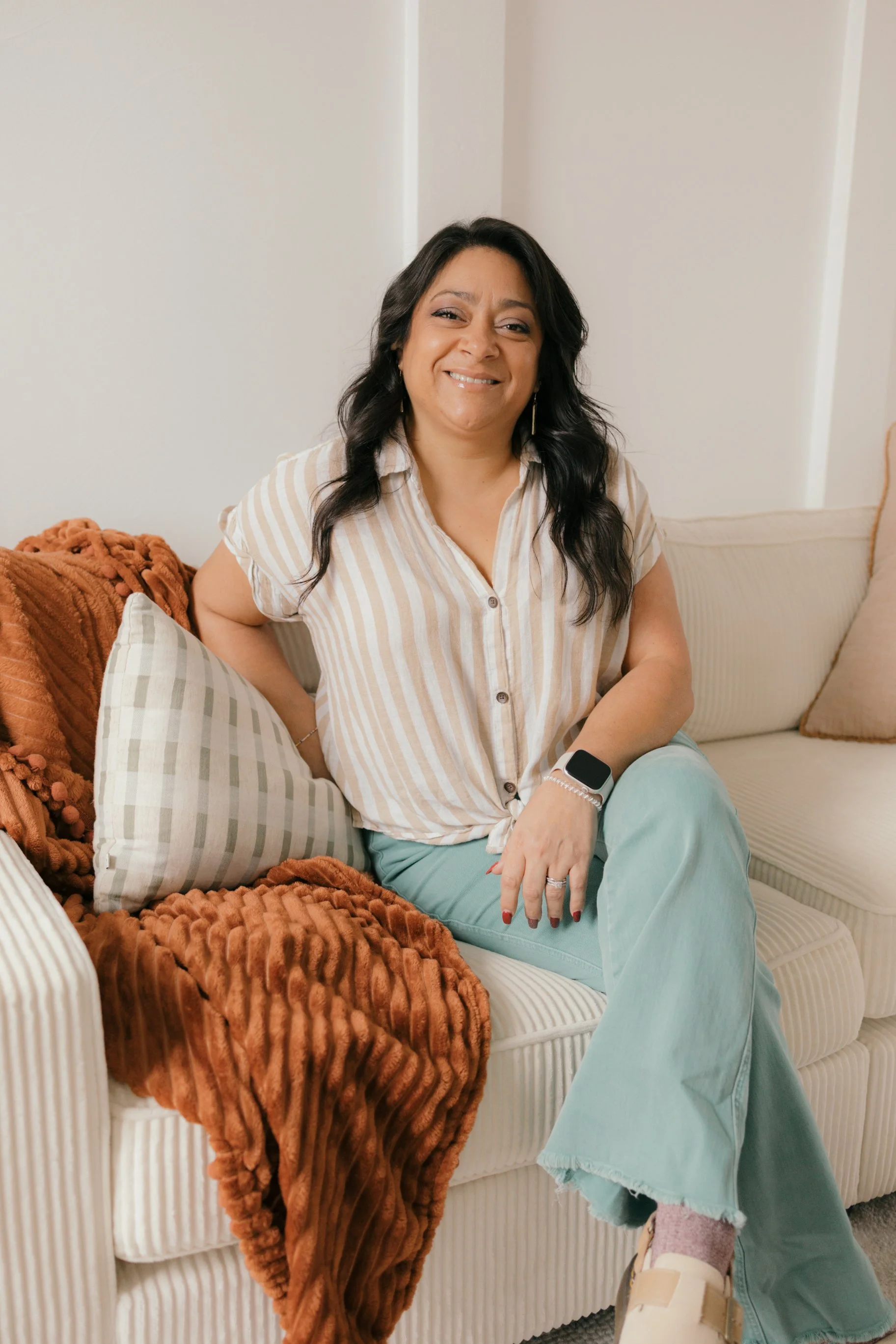 A woman with dark wavy hair smiling, sitting on a beige striped sofa, wearing a beige striped button-up shirt and light green pants, with a smartwatch on her wrist, and a brown plush blanket on the sofa.