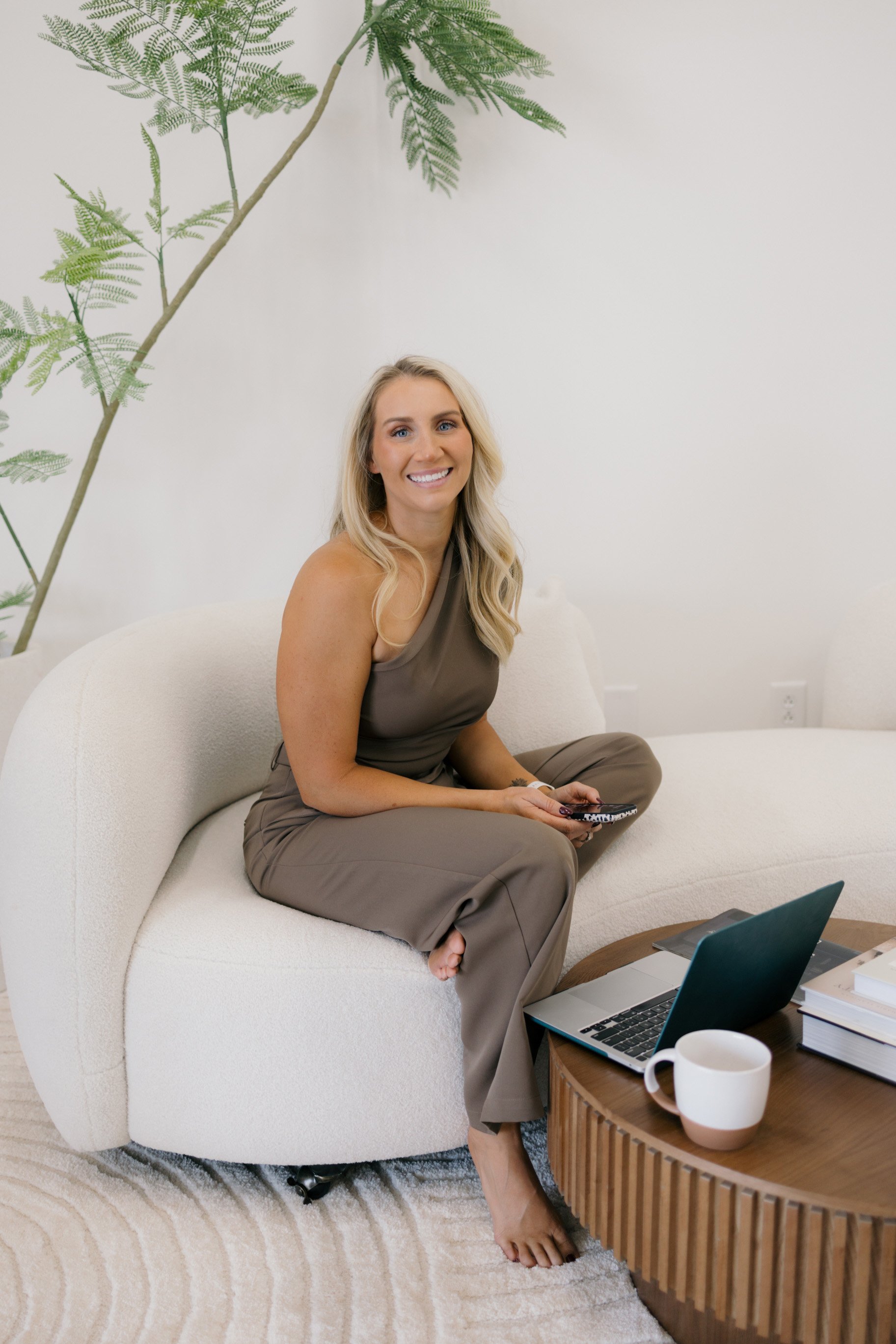 A woman with long blonde hair smiling and sitting on a white curved couch in a modern room. She is holding a remote control, dressed in a taupe top and matching pants, with one foot on the floor and the other leg crossed over her knee. In front of her is a wooden coffee table with an open laptop, a white mug, and some books. To her left is a large green houseplant against a plain white wall.