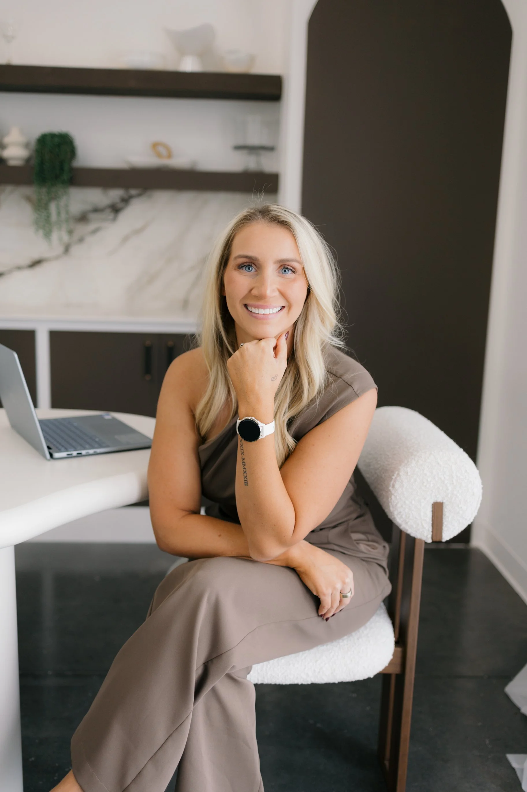 A woman with long blonde hair, blue eyes, and a smile, sitting in a room with a laptop on a white desk. She is wearing a taupe-colored outfit and a smartwatch on her left wrist. She is resting her chin on her right hand, sitting on a chair with a white fuzzy covering, with a black and white marble-patterned wall behind her.