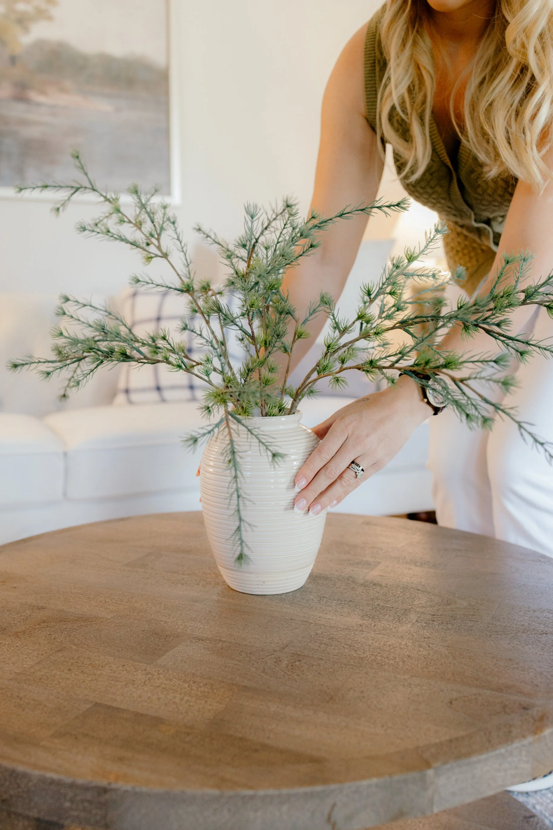 A woman with blonde, curly hair arranging a green plant in a white, textured ceramic vase on a round wooden table.