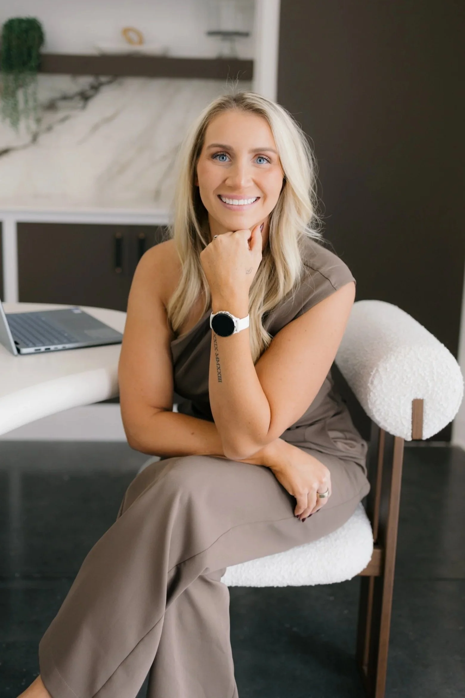 A smiling woman with long blonde hair, wearing a brown top, brown pants, a watch, and a ring, sitting on a cream-colored chair with a black wall and abstract artwork in the background.