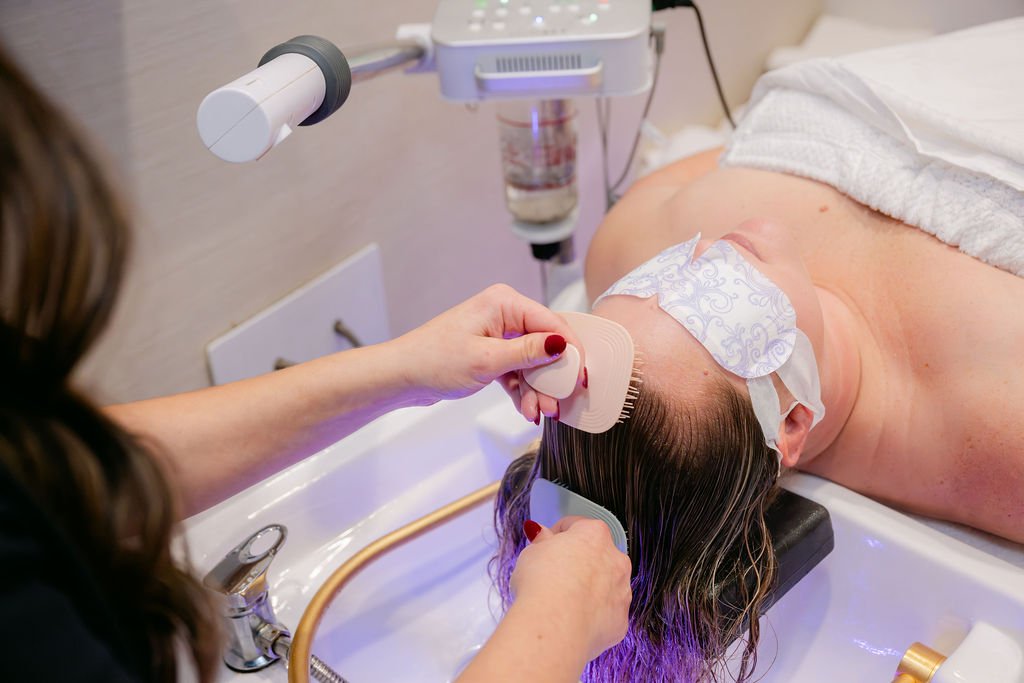A person receiving a hair treatment in a salon or spa, lying on a treatment bed with a towel over their eyes, while a stylist uses a brush and a comb to style or treat their wet hair.