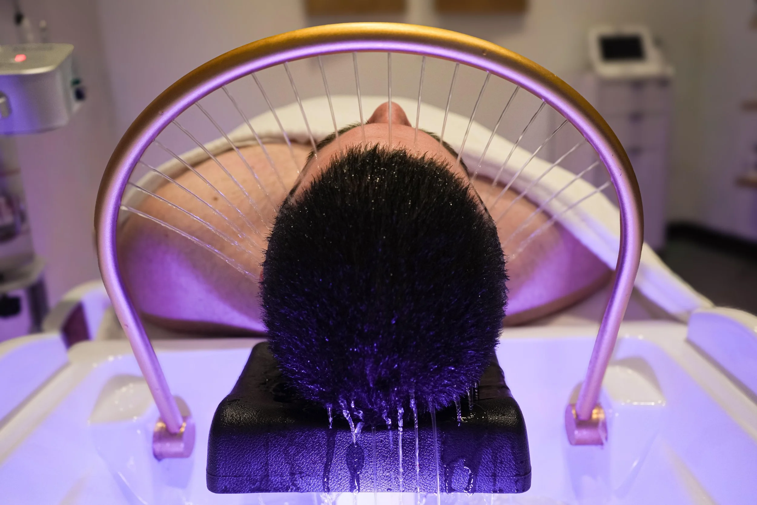 A person undergoing a facial treatment with a black brush and purple light in a spa or clinic environment.