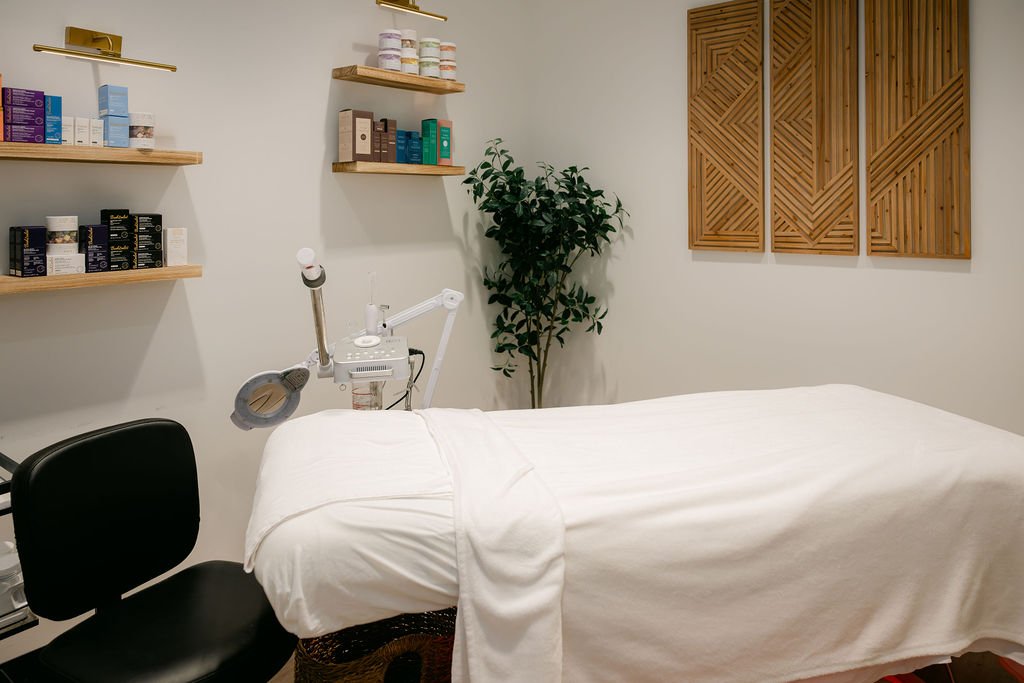 A massage or treatment room with a massage bed covered in white sheets, a black chair at the head of the bed, shelves with skincare products, a potted plant, and wooden wall decorations.