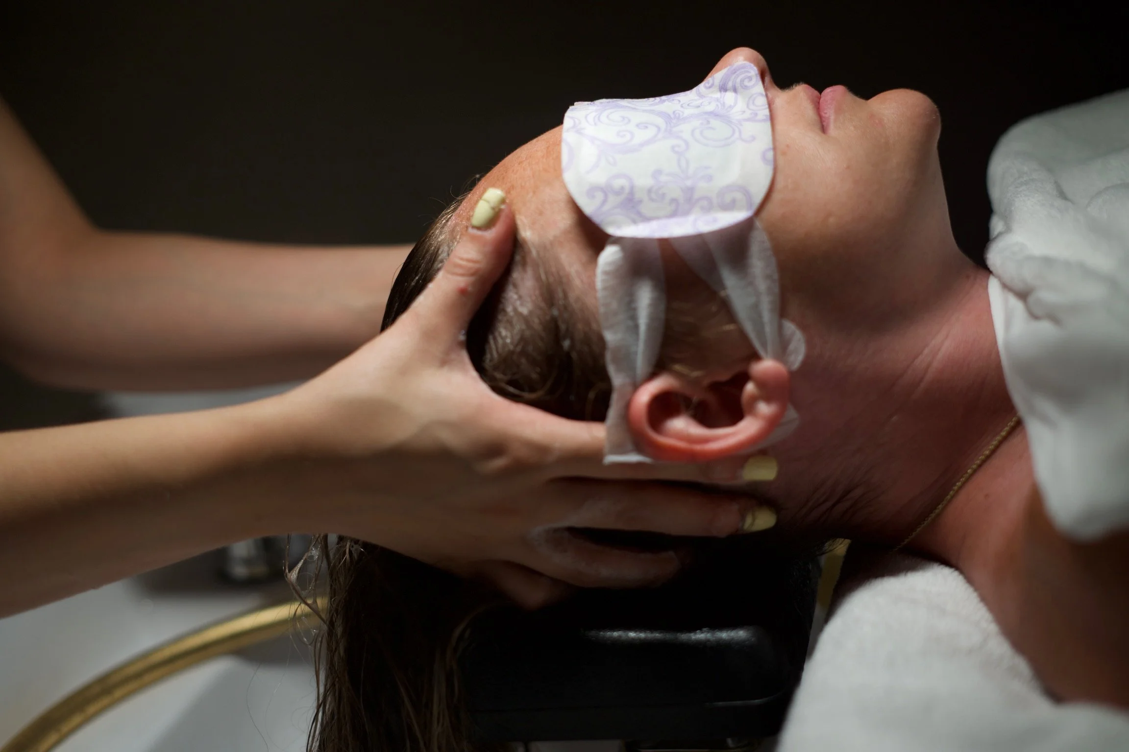 A woman receiving a hair treatment at a salon, lying back with hair being washed by a stylist.