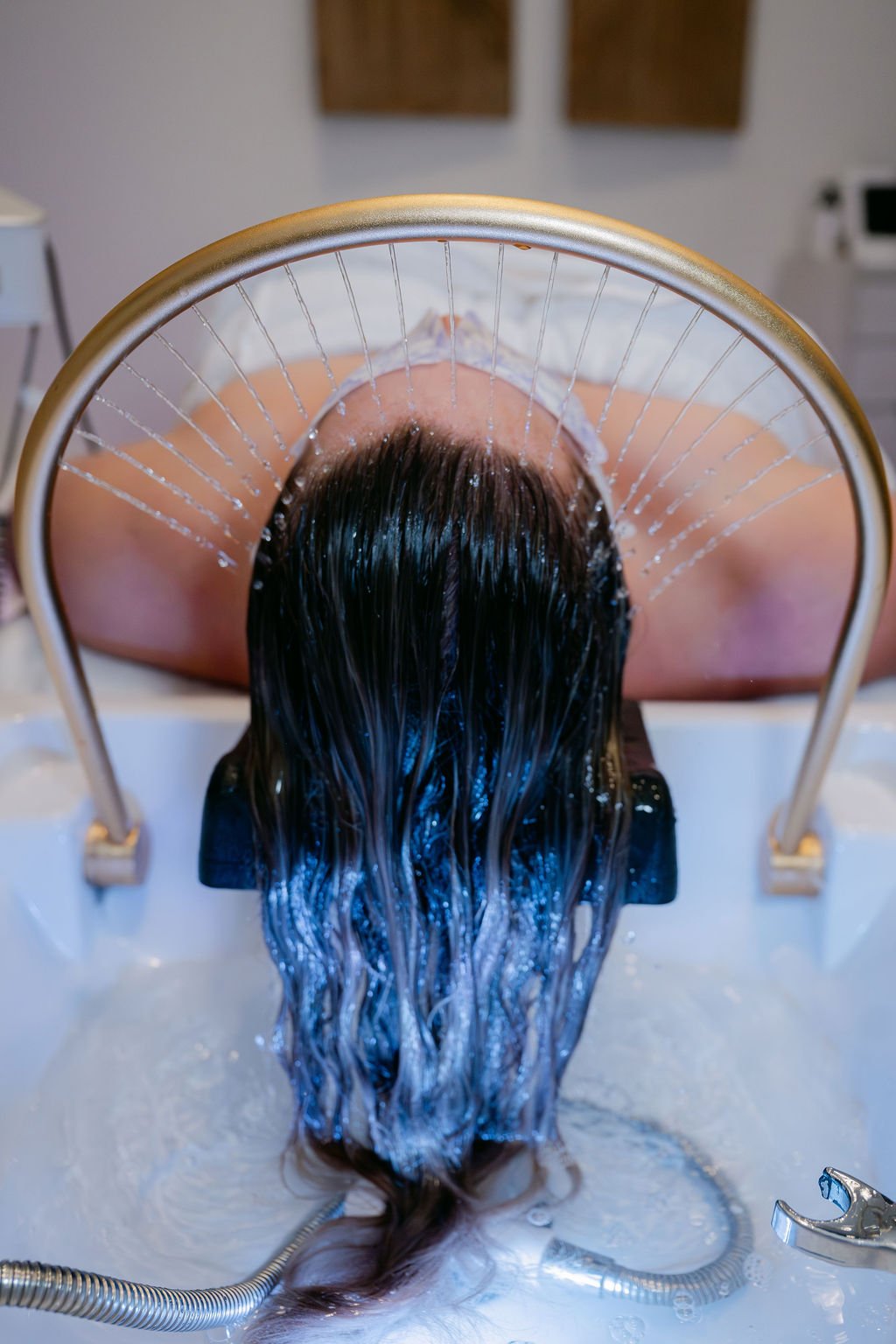 Person with long dark hair lying face down in a spa or massage bath, with water pouring over their head from a circular overhead shower.