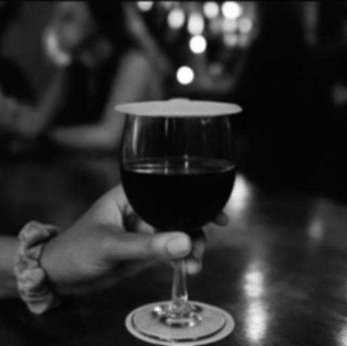 Person holding a glass of red wine with a coaster on top, in a dimly lit setting.