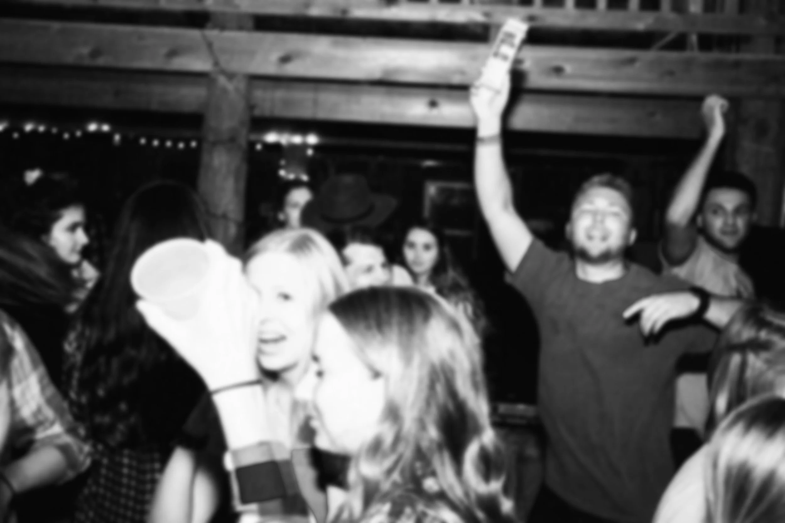 Group of people dancing and socializing at a party in a dimly lit setting with wooden beams, some with drinks in hand, smiling and enjoying the moment.