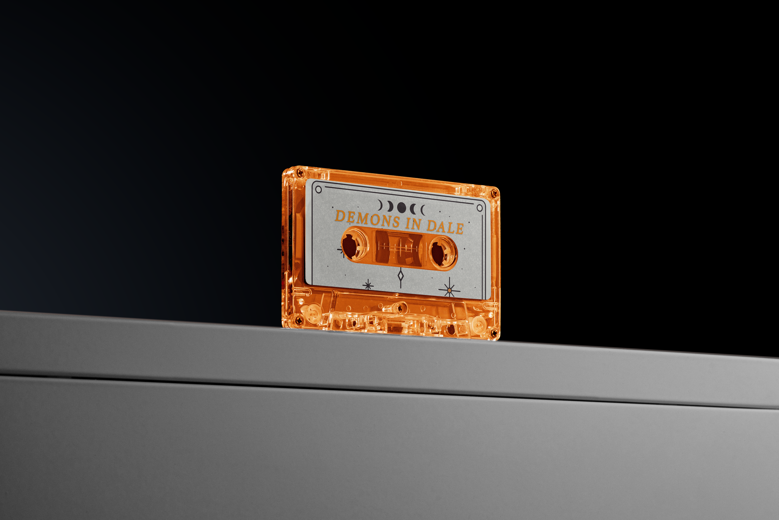 An orange cassette tape labeled "Demons in Dale" resting on a gray surface against a black background.