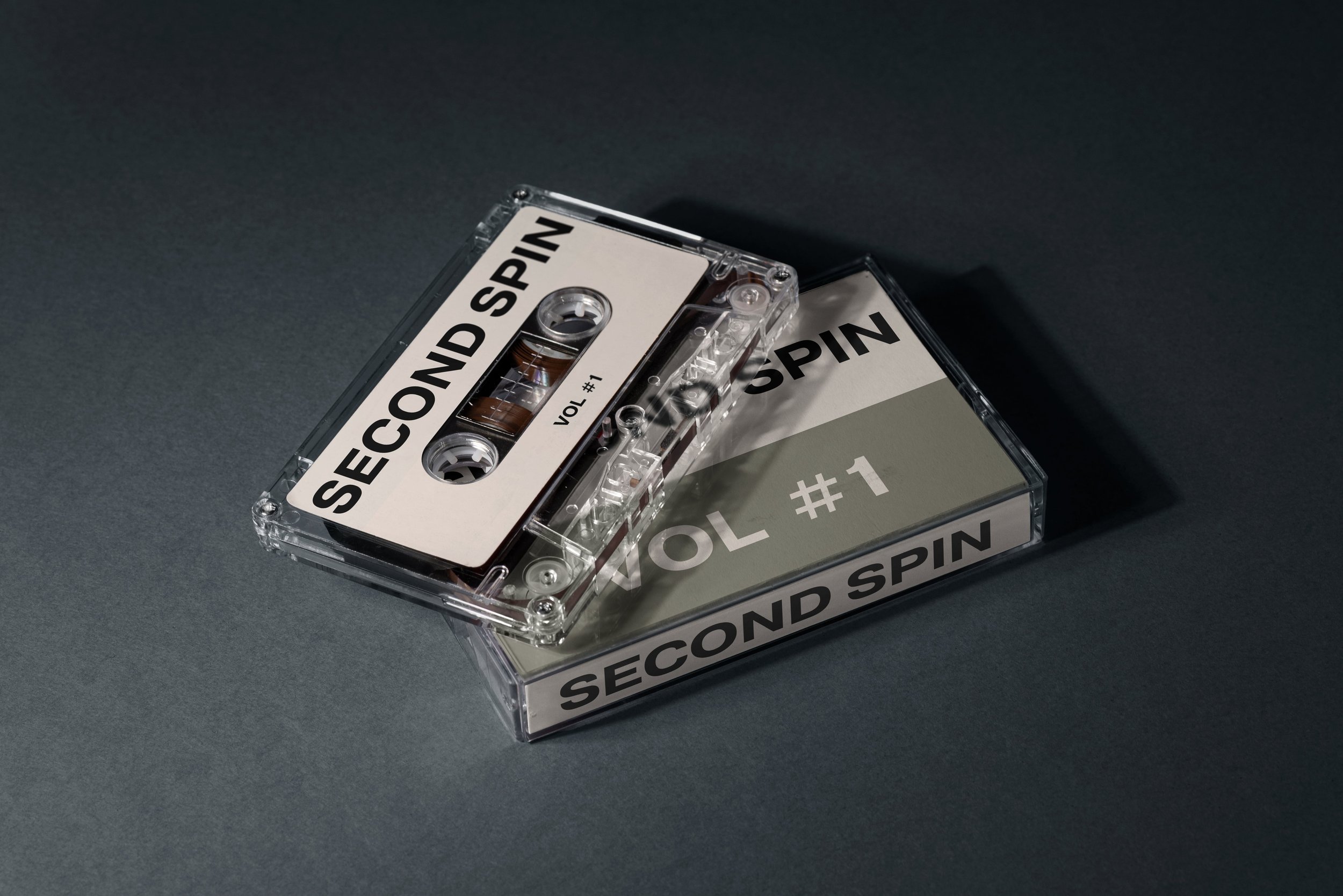 A clear cassette tape labeled 'SECOND SPIN' on the top, with 'VOL #1' on the label, resting on a dark surface.