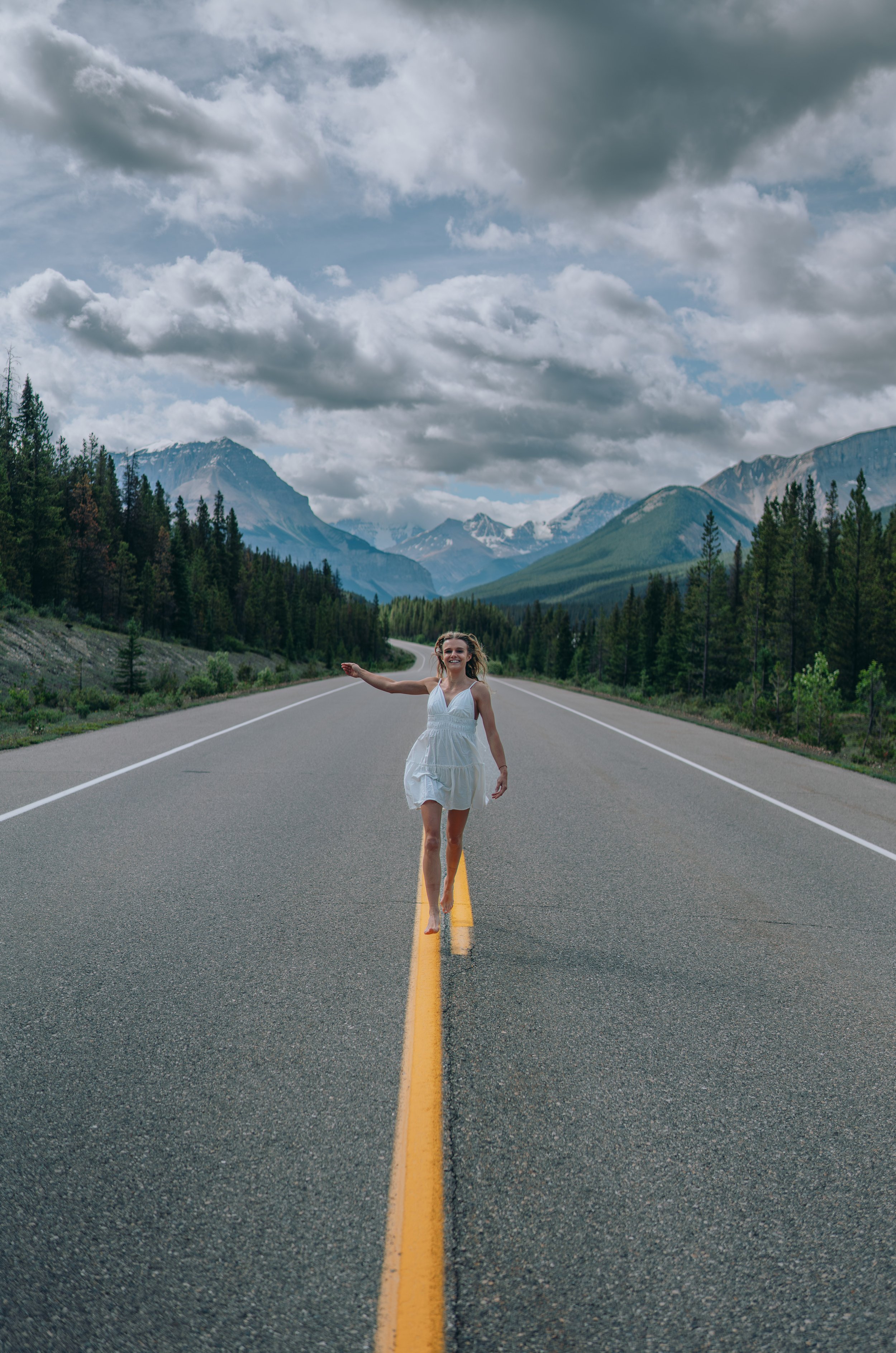 Chelsea Elwood, the Founder of Elwood Strategies, in a white dress walking barefoot on a mountain road with lush green trees and distant snow-capped mountains under a cloudy sky.