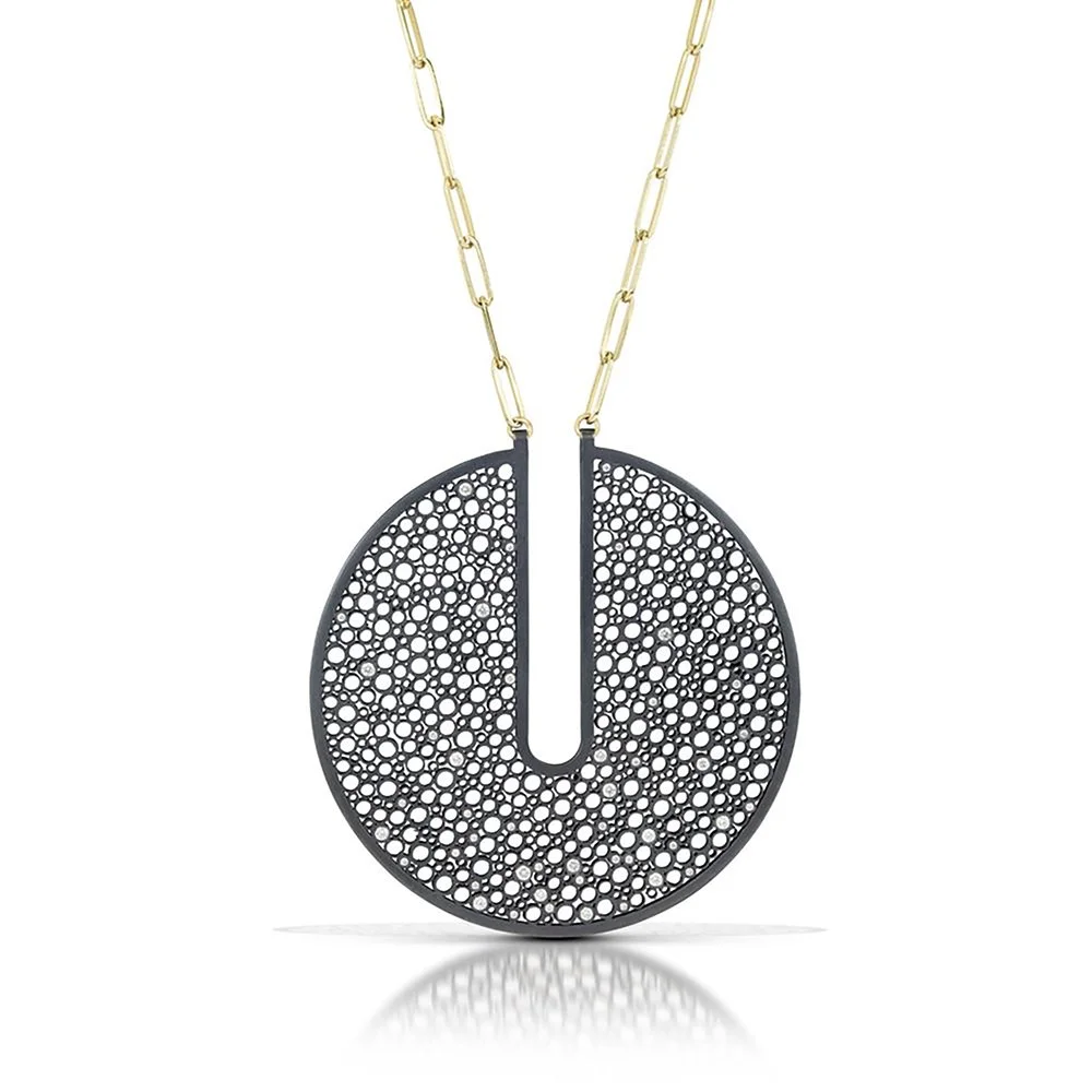 A modern necklace with a circular black pendant featuring a pattern of numerous small holes, hanging from a gold chain.