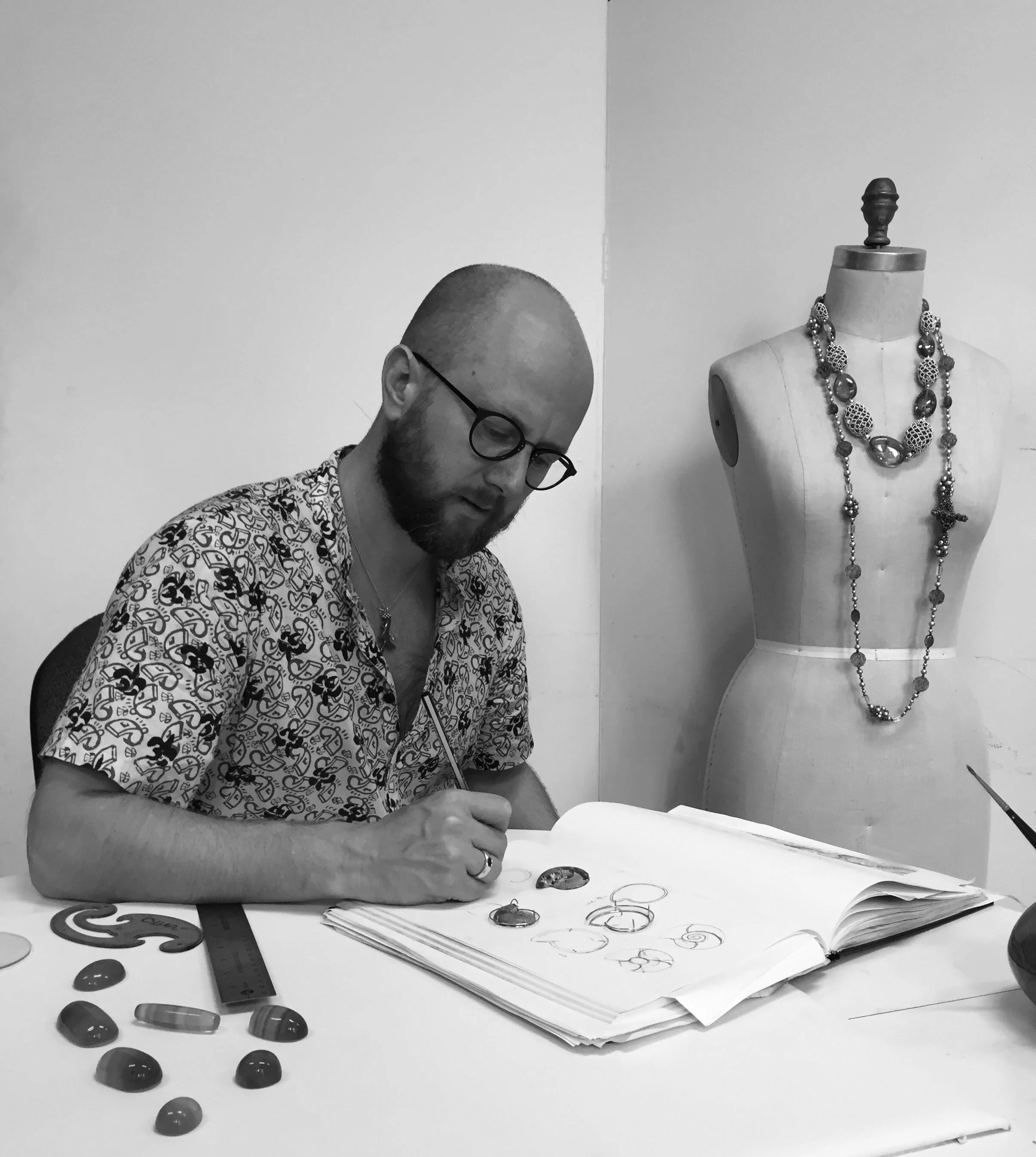 A man with glasses and a patterned shirt is sitting at a desk, working on jewelry sketches on a notebook. On the desk are various jewelry pieces, drawings, a ruler, and some loose gemstones. In the background, a mannequin displays layered necklaces.