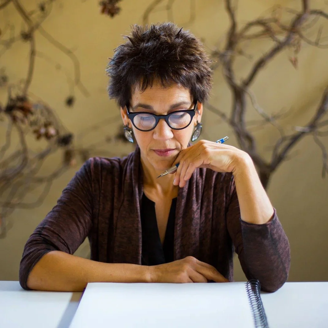 A woman with short dark hair, glasses, and earrings sitting at a table, looking down at a notebook with a pen in hand, in front of a wall with a vine pattern.