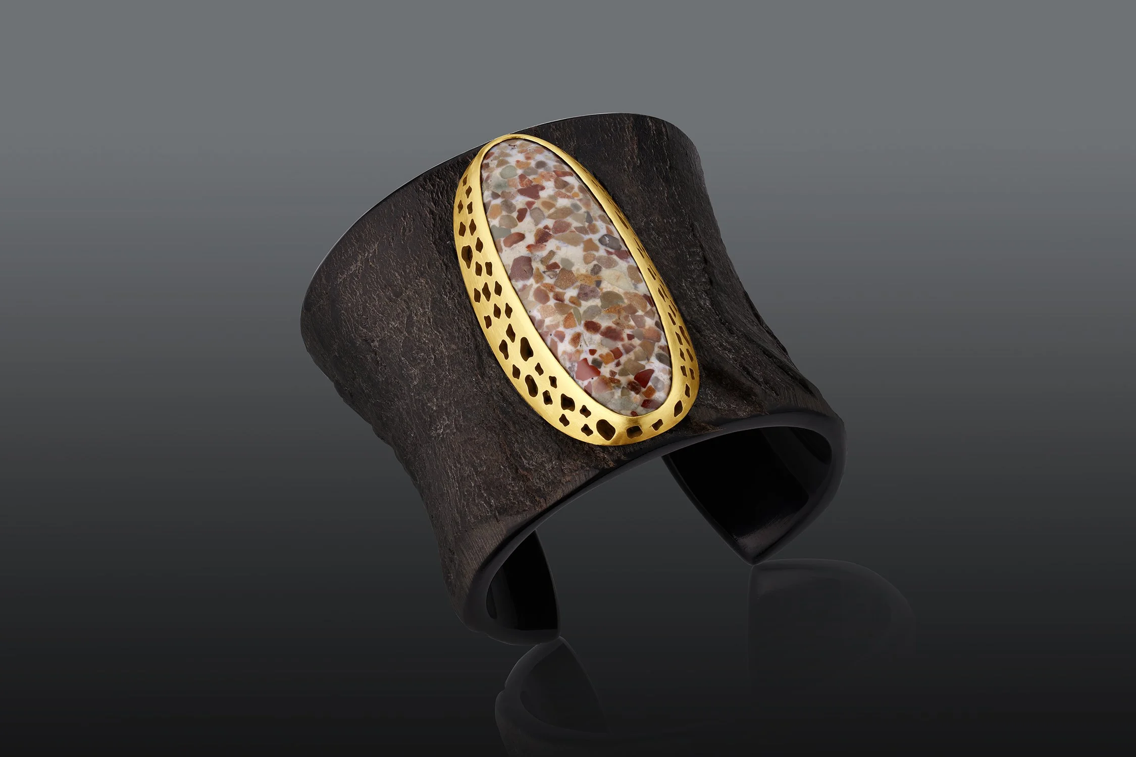 A black ring with a large oval gemstone featuring multicolored pebble-like fragments, set in gold, resting on a piece of dark textured wood against a gray background.