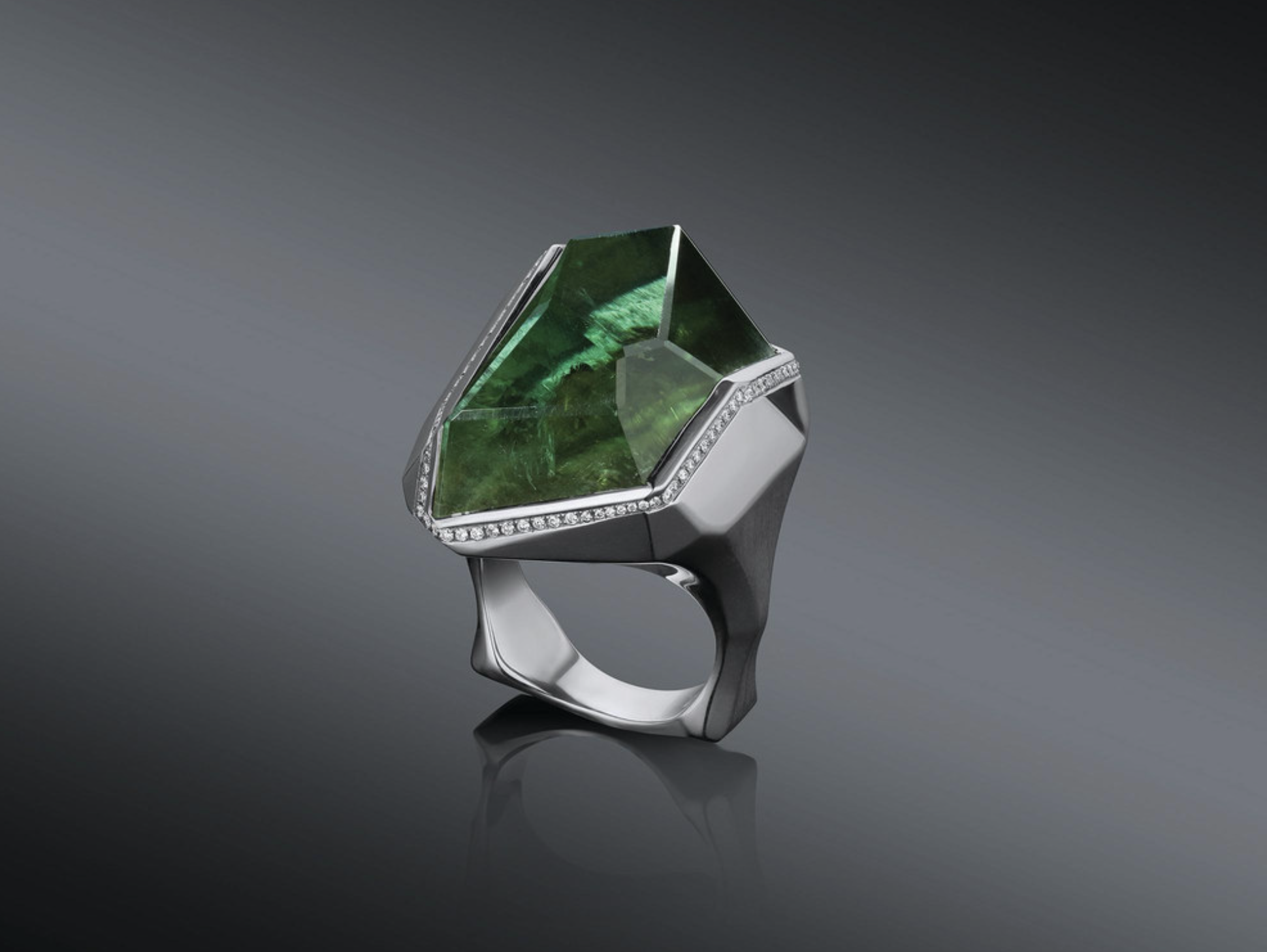 A silver ring with a large, irregularly shaped green gemstone and a border of small diamonds, showcased on a dark background.