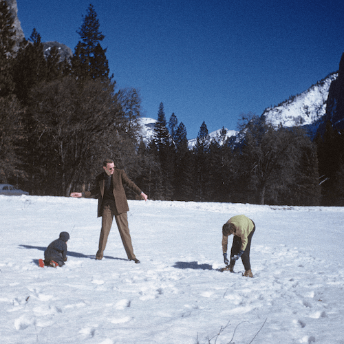 Three people playing in the snow in a mountain landscape with trees and snow-covered mountains in the background.