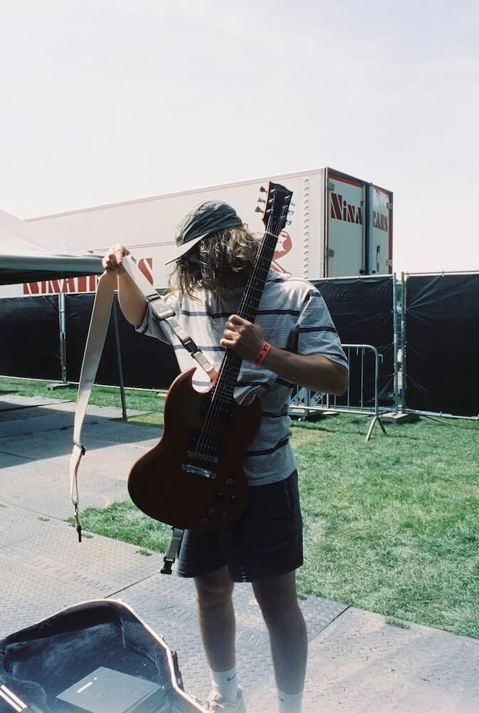 Divided Live - A person with long hair, wearing a cap, striped shirt, and shorts, playing an electric guitar at an outdoor event with grass, fencing, and trailers in the background.