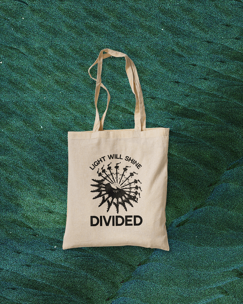 Beige tote bag with black text and design on a green textured background. The text reads 'Light Will Shine Divided' and the design features a spiraling pattern of arrows or rays.