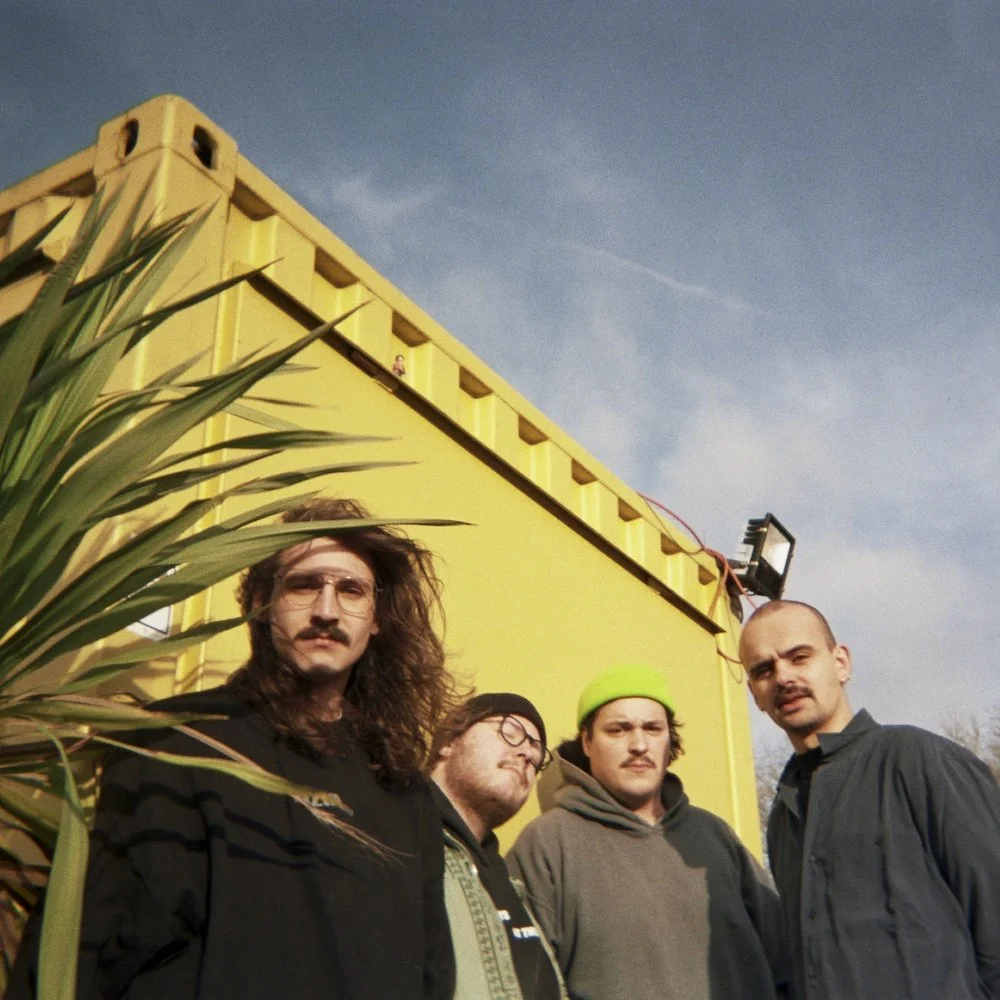 Divided Press - Four men with serious expressions standing outdoors in front of a yellow container or building, with a plant partially obscuring the left side of the image. The sky is partly cloudy.