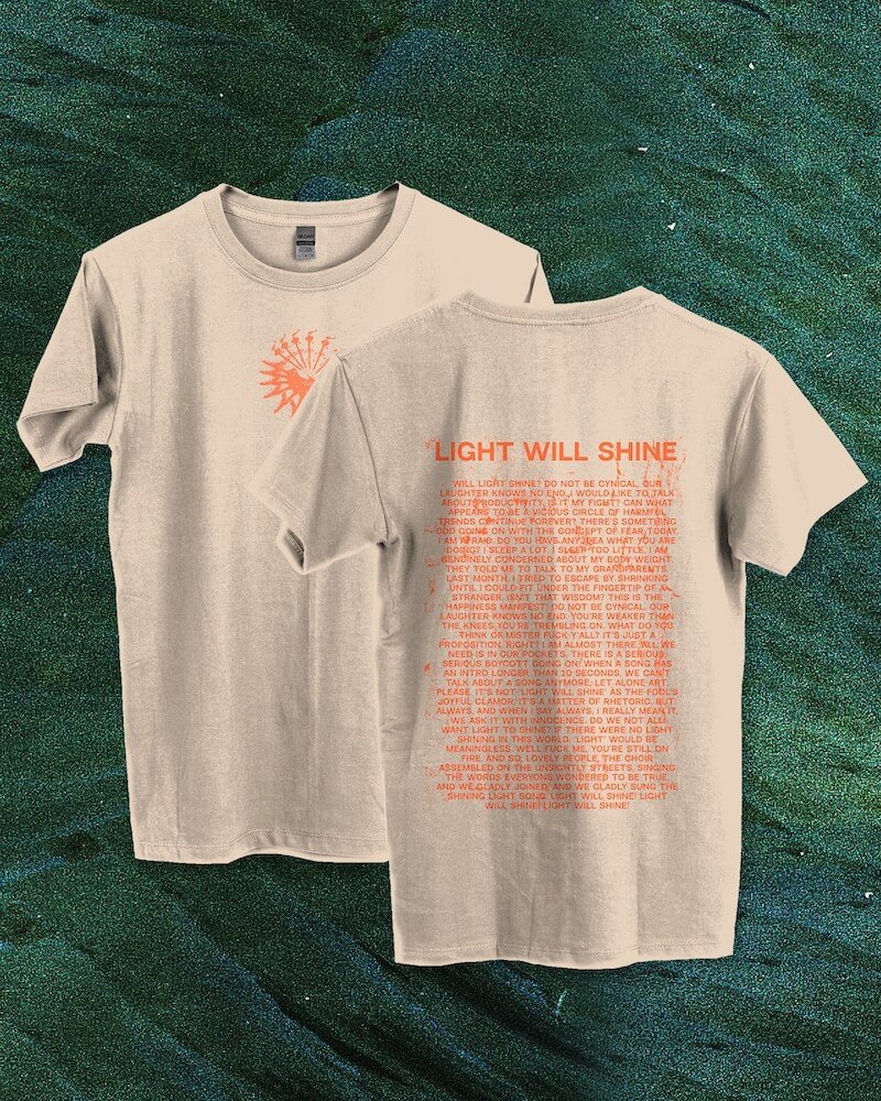 Two beige T-shirts with orange text and design, one displaying a sunburst graphic with text "LIGHT WILL SHINE" and the other with a block of orange text on the back, laid on a dark green textured background.