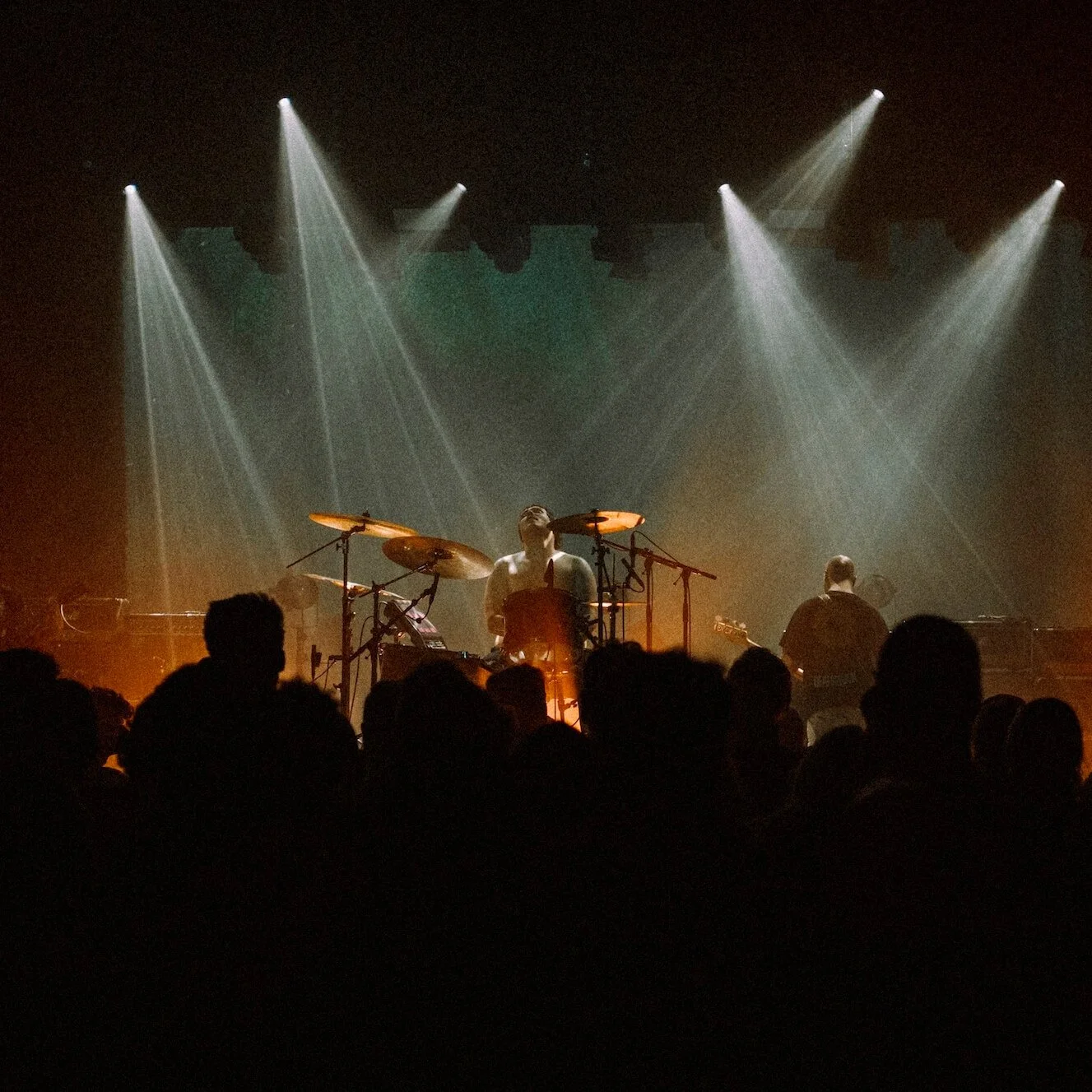 Divided Live - A concert scene with a drummer on stage illuminated by spotlights, with an audience visible in the foreground.