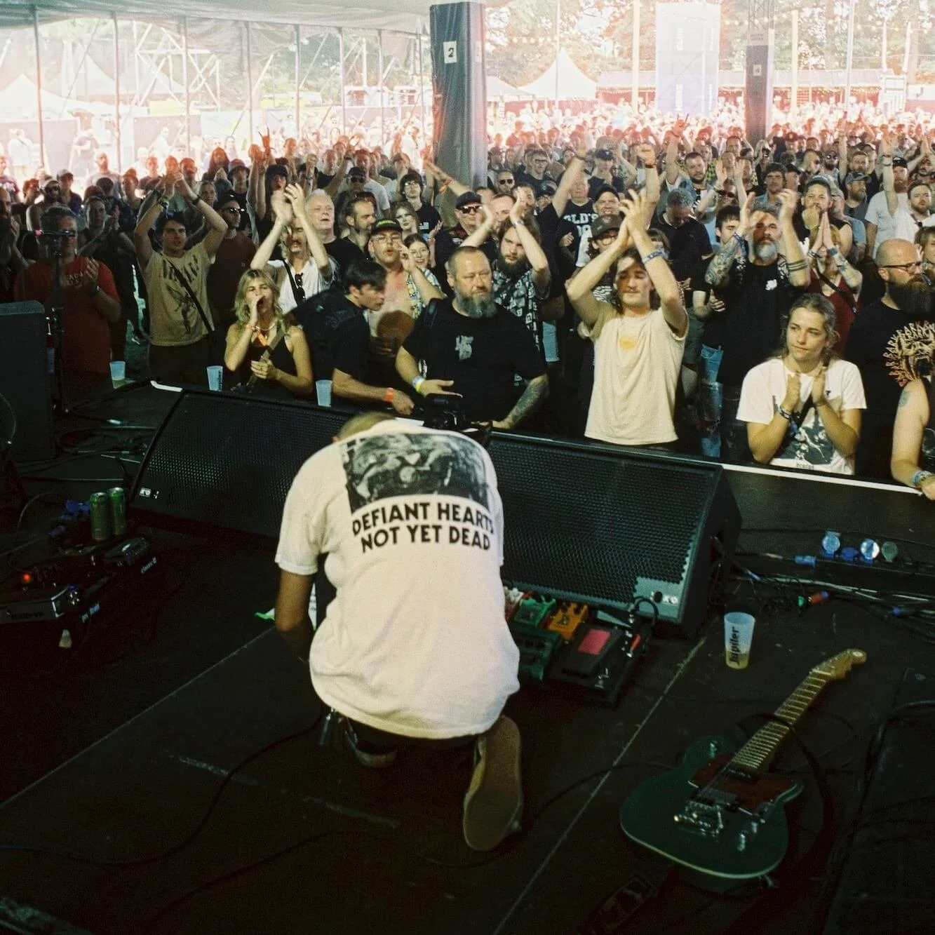 Divided Live - A musician kneeling on stage adjusting equipment while an audience cheers and claps inside a large tent at a concert.