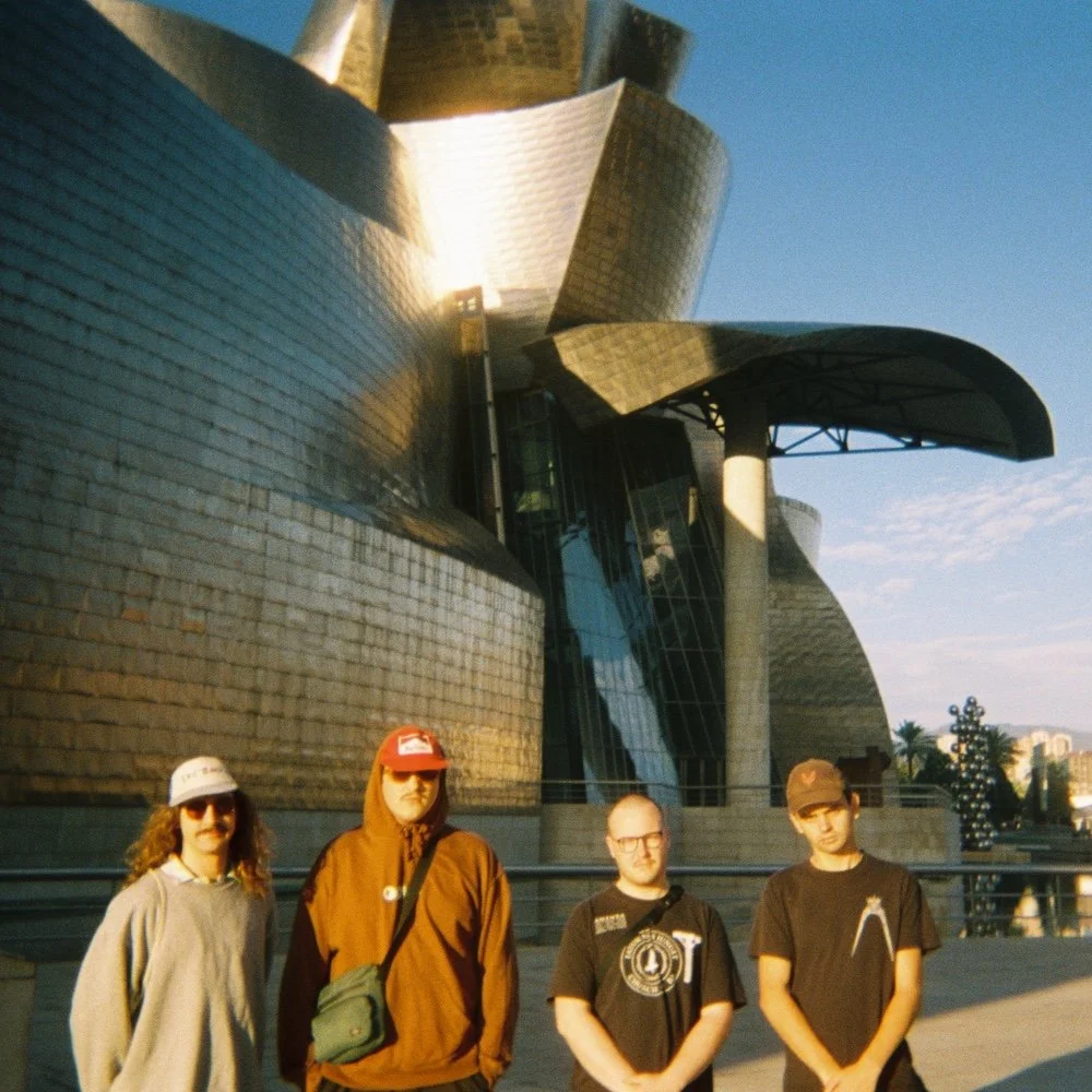 Divided Press - Four young men stand in front of the Walt Disney Concert Hall in Los Angeles, California, during daylight. The building has a modern, metallic, curvilinear design.