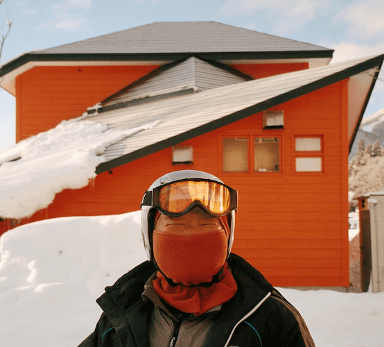 Person in winter gear wearing goggles and a face mask standing outside in front of a snow-covered orange house with snow piled on the roof.