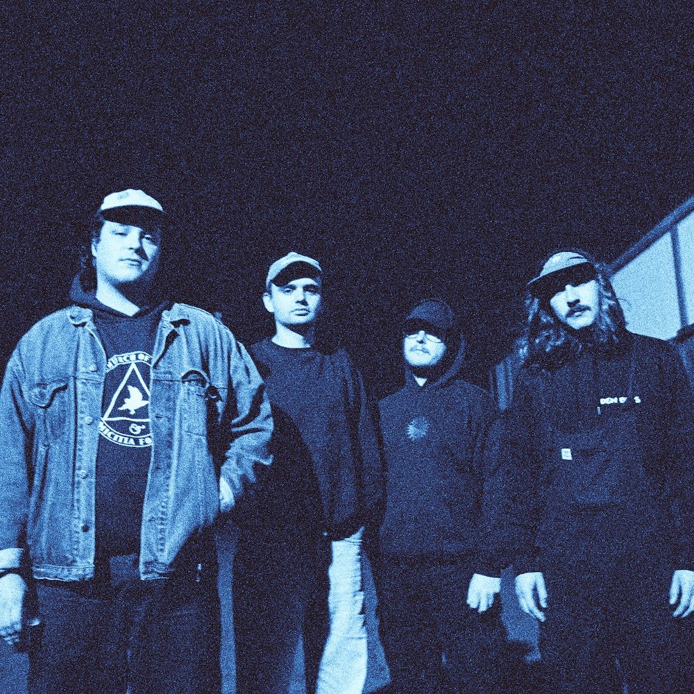 Divided Press - Four young people standing outdoors at night, wearing dark clothing and hats, with a mostly dark sky above.