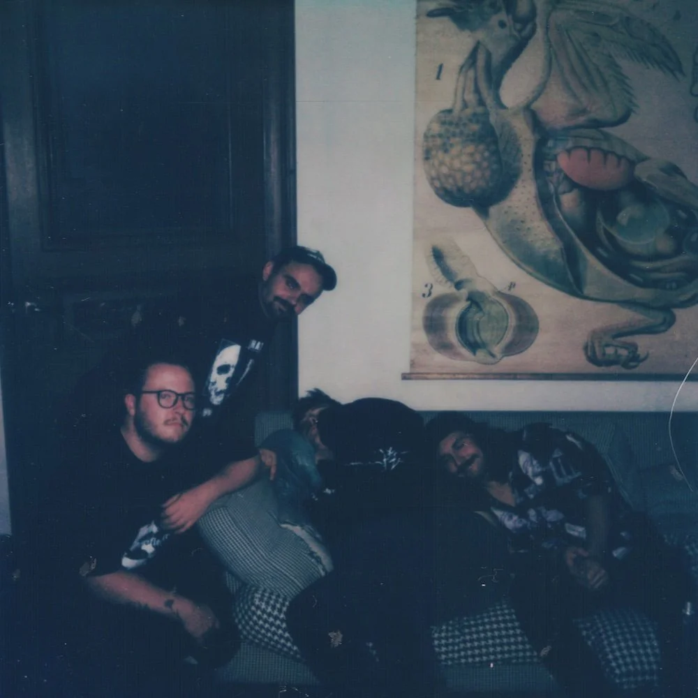 Divided Press - Four young men in a dimly lit room with a anatomical poster of the human heart on the wall.