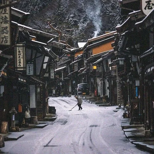 Snow-covered street in a traditional Japanese village with shops, a person shoveling snow, and smoke rising from a chimney.