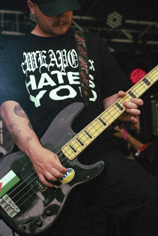 Man wearing a black shirt and cap playing an electric bass guitar with a sticker saying 'UAA' and a colorful strap, in a dark setting.