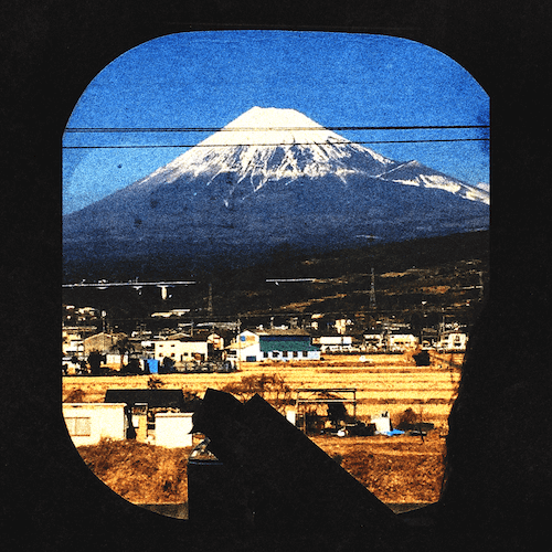 View of Mount Fuji through a train window with a small town in the foreground and power lines overhead.