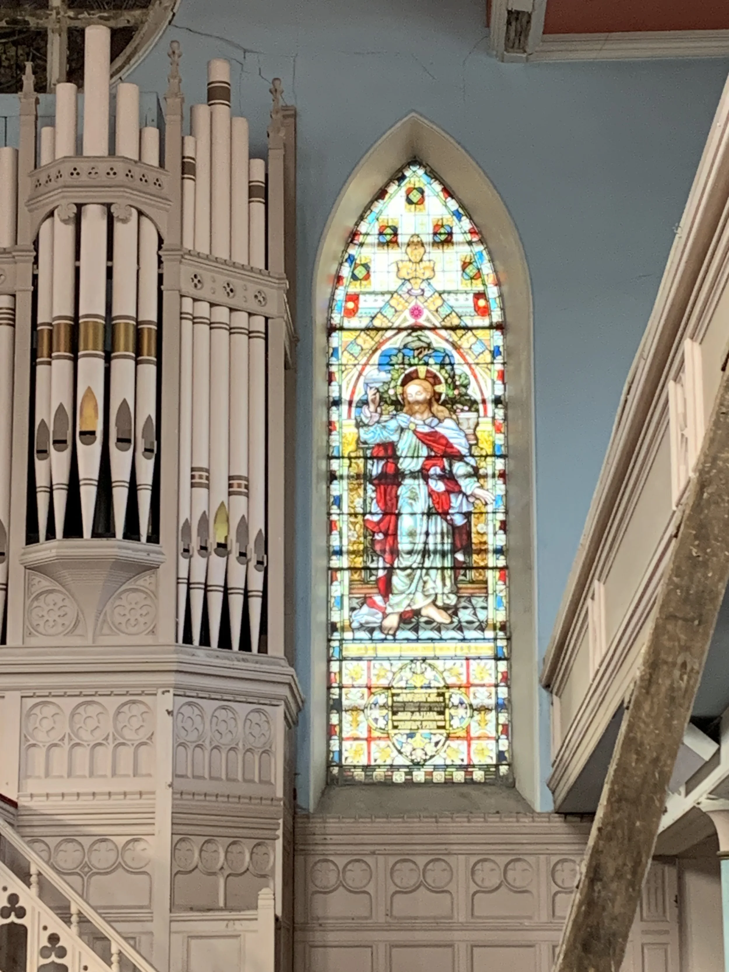 Colorful stained glass window in a church depicting Jesus Christ with a halo, holding a staff, surrounded by intricate patterns, with a pipe organ to the left.