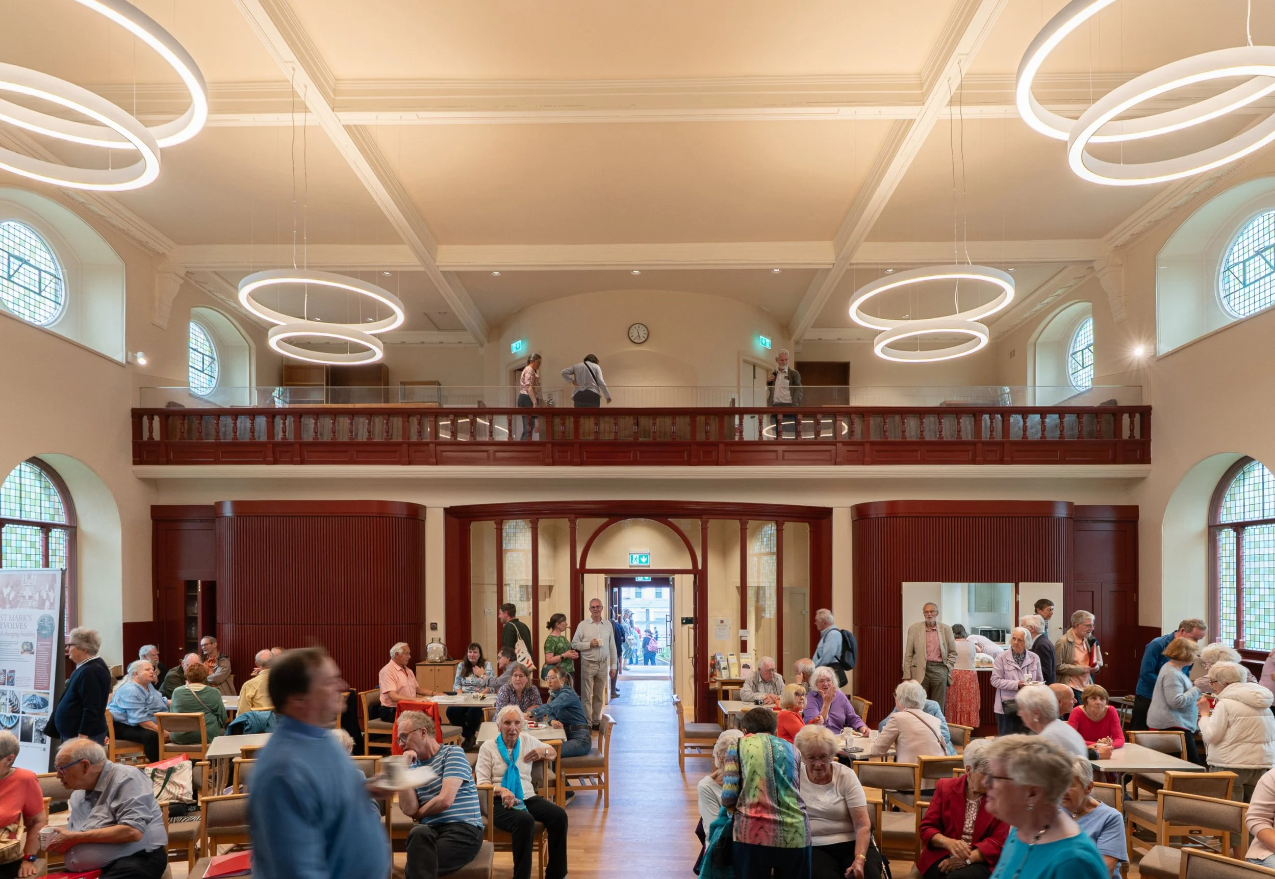 A crowded indoor space with many elderly people sitting at tables and socializing. There are people standing and walking around, and a few individuals are on a balcony overlooking the lower level. The room has high ceilings, large arched windows, and modern circular pendant lights.