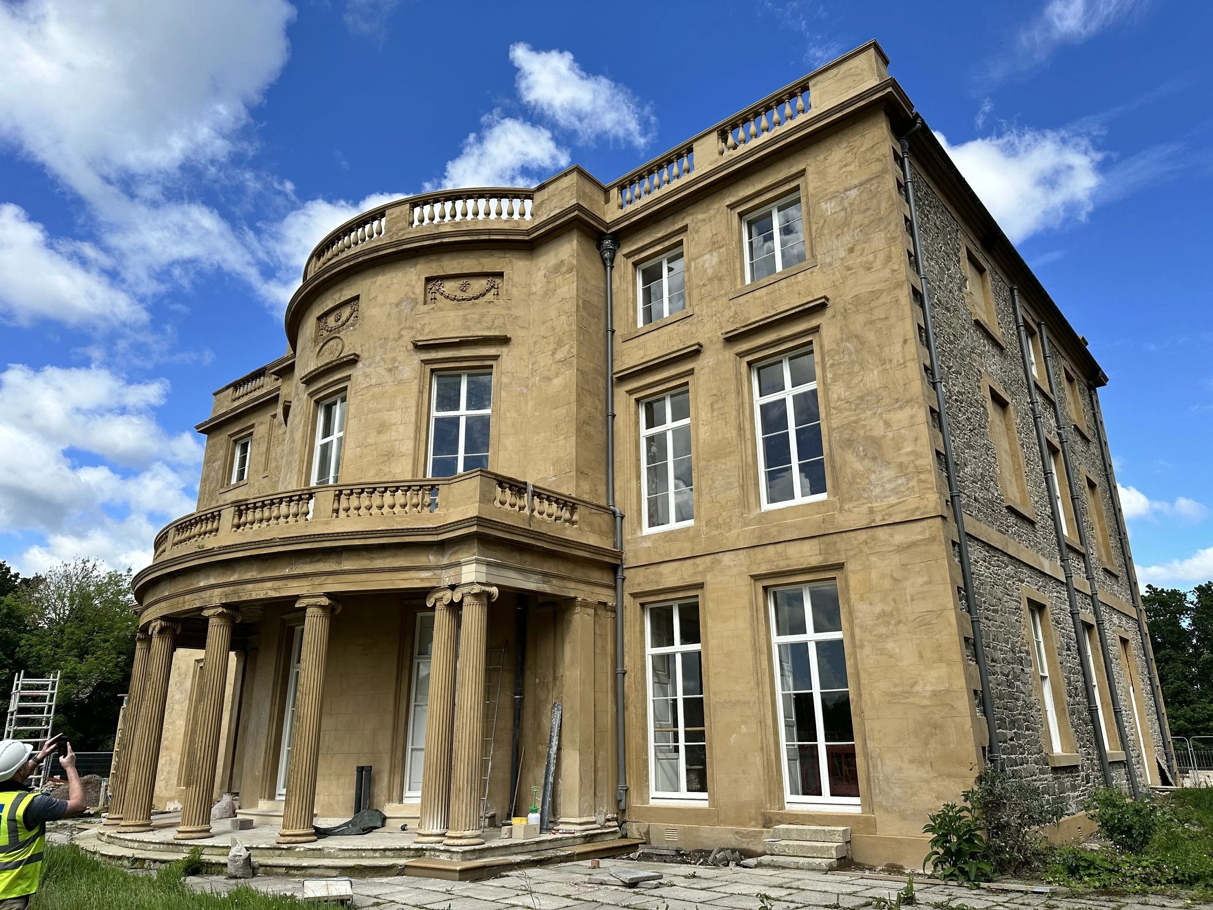 Construction site of a large, historic-style mansion with beige stone facade, tall white-framed windows, and Corinthian columns at the entrance. Part of the building exposed with stonework visible. Worker in safety vest and helmet taking a photo, blue sky with clouds overhead.