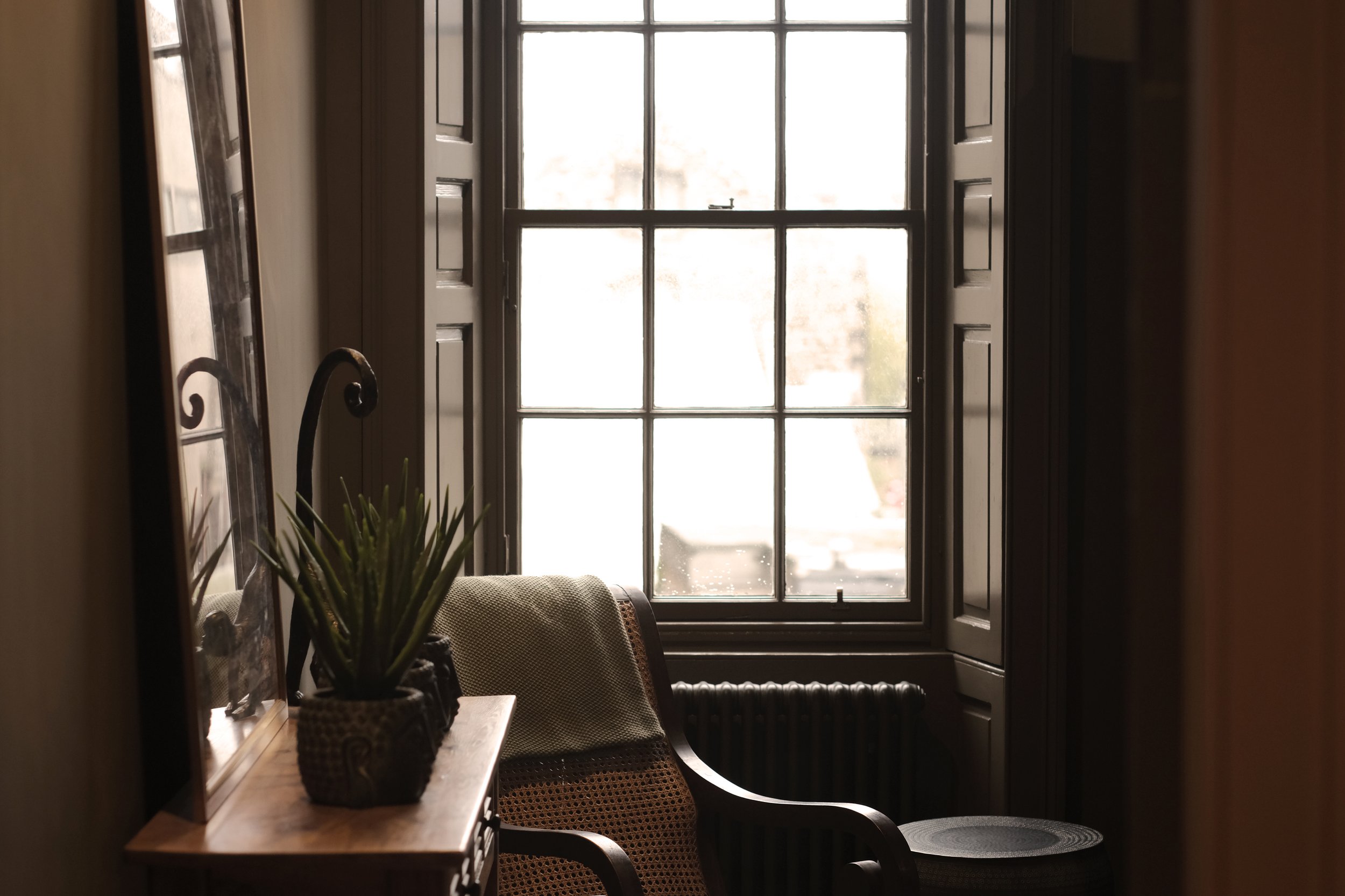 A cozy interior scene with a window, a chair, a side table with a potted plant, and a mirror, bathed in natural light.