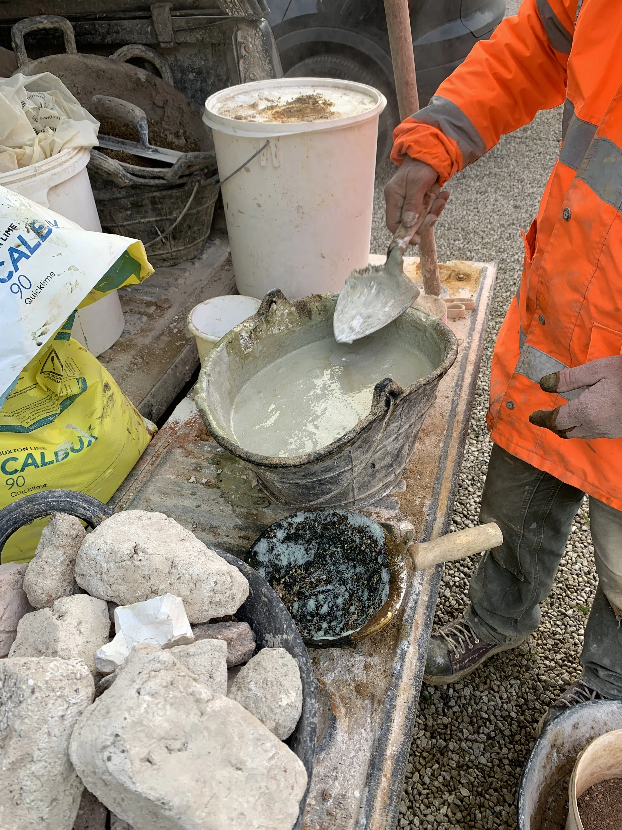 A construction worker in an orange safety jacket and muddy gloves stirs a mixture in a metal bucket at a construction site. Various construction materials and tools, including bags of quicklime, rocks, and buckets, are visible on the work surface.