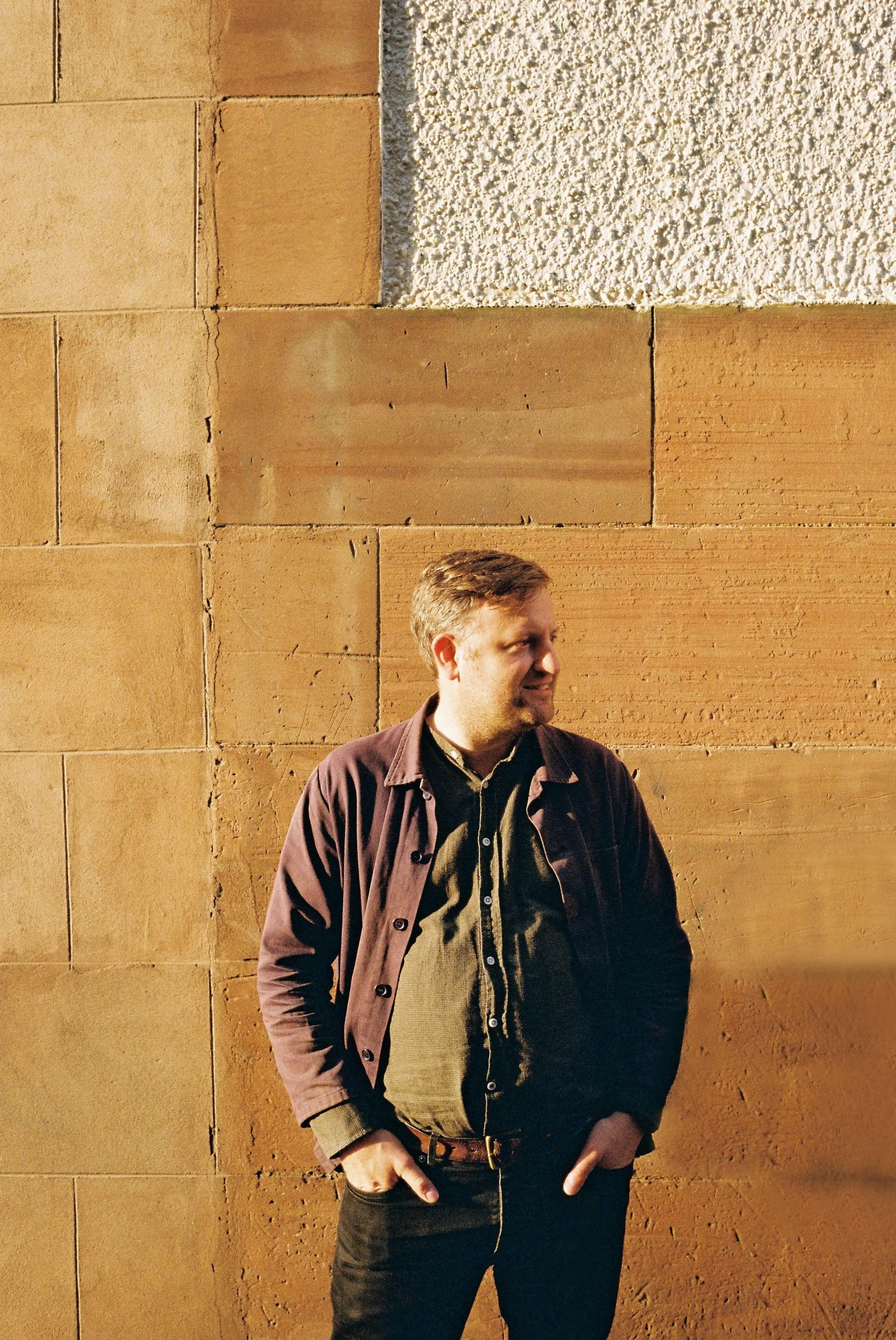 A man with light brown hair and a beard stands against a tan and white stone wall at night, wearing a brown jacket over a green shirt with hands in pockets.