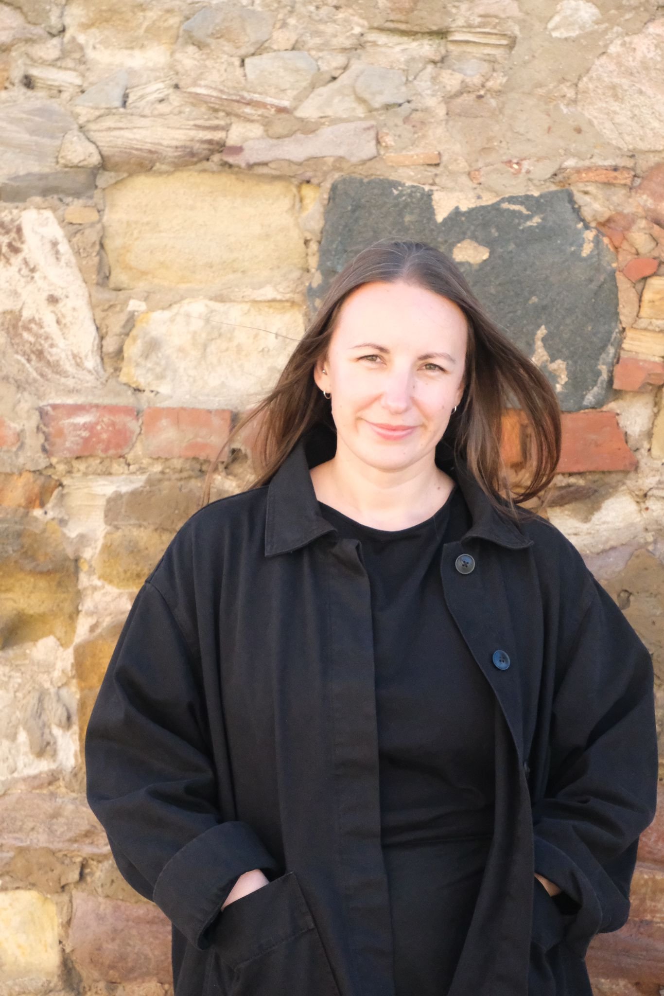 A woman with long brown hair wearing a black jacket standing in front of a rustic stone wall, smiling slightly.